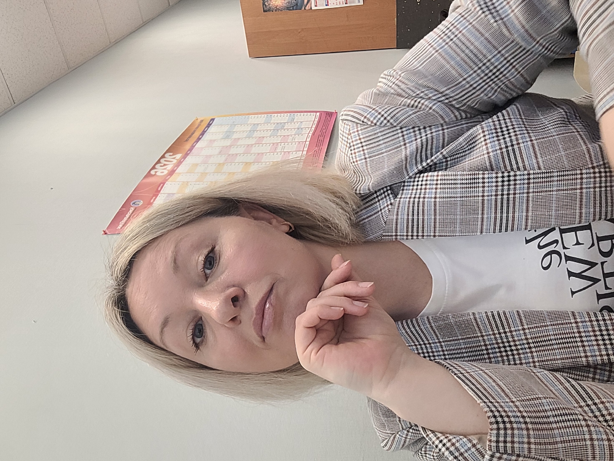 A woman with short blonde hair and blue eyes is posing for a selfie. She is wearing a white t-shirt with black lettering and a plaid blazer. Her hand is resting under her chin in a thoughtful gesture. Behind her, there is a wall with a calendar displaying the year "2023" visible. There is also what appears to be a wooden cabinet above the calendar.
The location cannot be determined based on the visual information provided. There is no indication of the city of Izhevsk, Russia in this image.