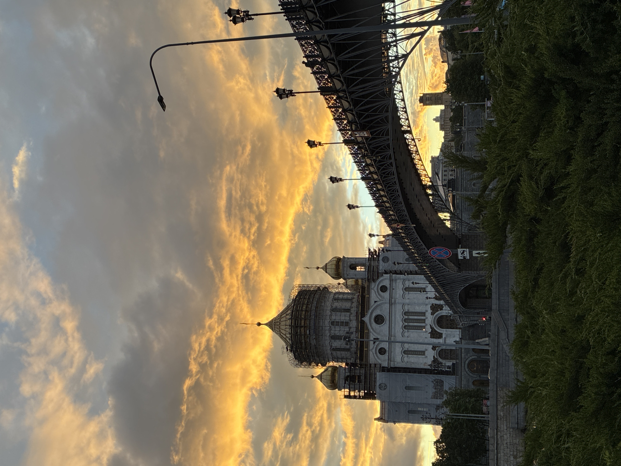 The image shows the Cathedral of Christ the Savior in Moscow, Russia, from a low angle, possibly street level. The cathedral is partially obscured by a bridge overhead. The sky is filled with clouds illuminated by a sunset. Scaffolding surrounds parts of the cathedral, suggesting ongoing construction or restoration work. Lush green bushes are visible on the right. Streetlights are attached to the bridge. A traffic sign is visible on the bridge.