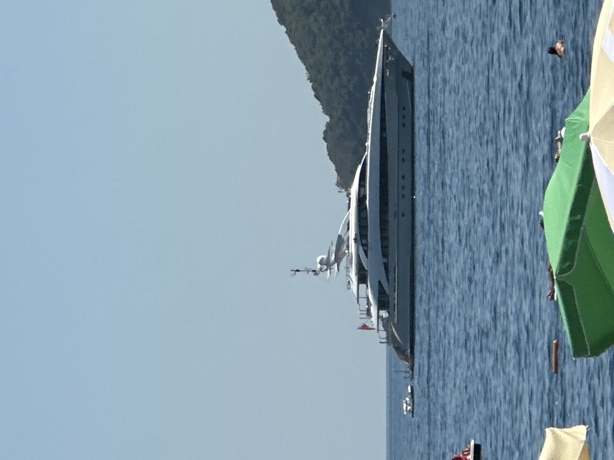 A wide-angle, eye-level shot captures a large white yacht with multiple decks anchored in the calm, blue waters off the coast of Budva, Montenegro. The yacht is positioned in the middle of the frame, extending horizontally across the image. A smaller white boat is visible to the left of the yacht, closer to the shore. In the background, a lush, green, tree-covered hill rises steeply from the water's edge. The sky is clear and a pale blue. In the foreground, partially visible beach umbrellas, one yellow and white and another green, suggest the presence of a beach or swimming area. Two people are in the water in the lower right of the frame, one of whom is swimming. The overall scene depicts a sunny day with leisure activities on the water.