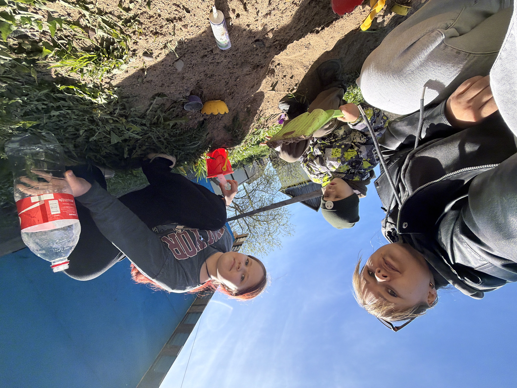 The image depicts a scene in Чумна, Russia, showing three individuals outdoors on a sunny day. A woman with auburn hair, wearing a gray t-shirt, holds a plastic Coca-Cola bottle. Beside her, a child in a camo jacket and a dark beanie holds a green shovel, seemingly playing in the dirt. Another woman in a black leather jacket and blue jeans looks at the camera. Various items such as dirt, a sealant tube, and sand toys, including a red bucket and yellow molds are scattered on the ground. To the left, there is vegetation and a blue structure of some sort. The clear blue sky is visible in the background.