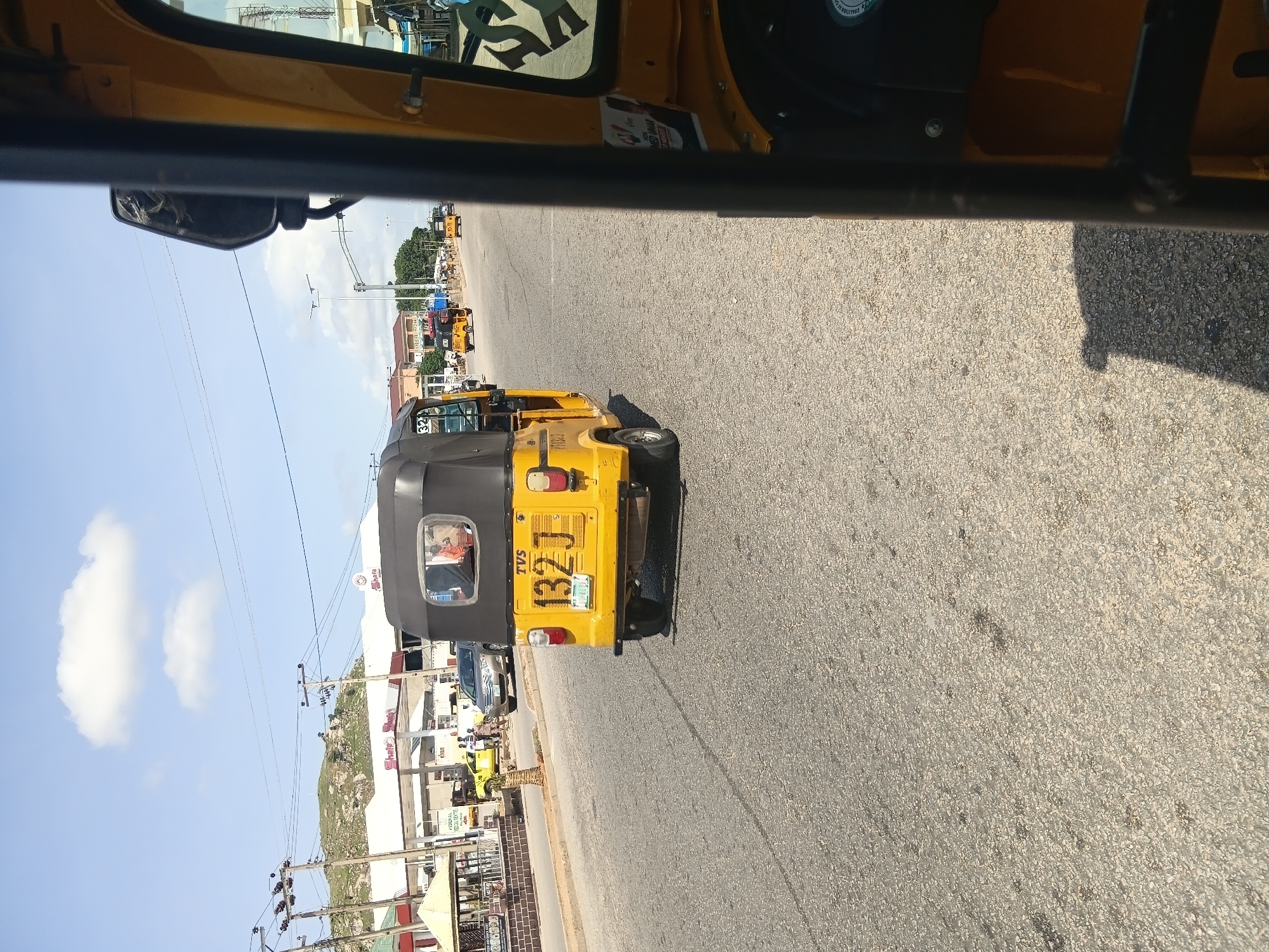 A yellow three-wheeled motorized rickshaw, identified as a TVS model with license plate number 132 J, is driving on an asphalt road in Jalingo, Nigeria. The rickshaw has a black canopy and two passengers are visible inside. The scene takes place during the day under a clear blue sky with scattered clouds. On the left side of the road, there is a gas station with a white canopy structure and a few other vehicles, including a silver car and another yellow rickshaw. Buildings and trees are visible in the background, and power lines stretch across the sky. The perspective is from the interior of another yellow rickshaw, looking forward.
