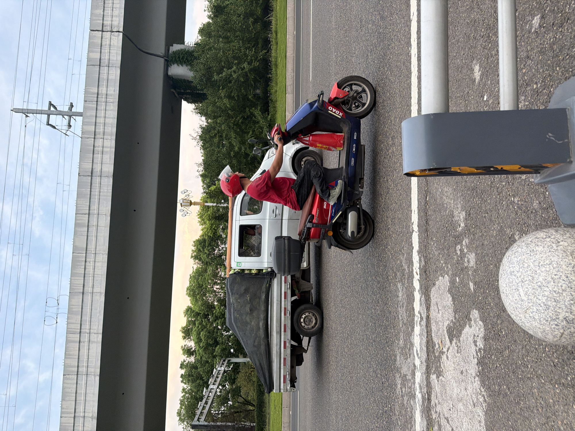 A man wearing a red helmet and a red t-shirt rides a red and blue electric scooter on a paved road in Jiangjin, China. The scooter has a red bag hanging from the front. Behind the scooter, a flatbed truck is driving, carrying a large black tarp. The truck is silver and has a white cab. In the background, trees and an elevated highway are visible. The sky is cloudy. A concrete barrier with reflective yellow and black stripes is in the foreground on the right side of the image.