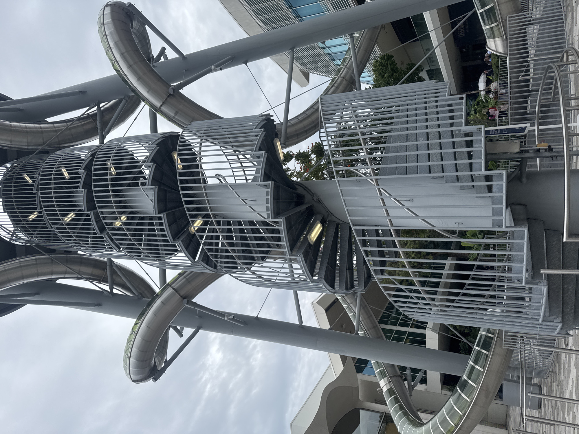 The image captures a unique architectural structure in the city of Aventura, Florida, United States. This structure features a combination of a spiral staircase and a slide system. The staircase is constructed with a gray metal frame, enclosed by a cage-like structure. Lights are integrated into the staircase's design, providing illumination.
The slide system consists of a series of curving tubes, also made of metal, which appear to wind around the staircase. Sections of the slides are transparent, allowing a view from within.
The structure is situated outdoors, against a backdrop of modern buildings with glass and metal facades. Some greenery, including trees and plants, are visible in the vicinity of the structure. People are visible on the ground level near the structure.