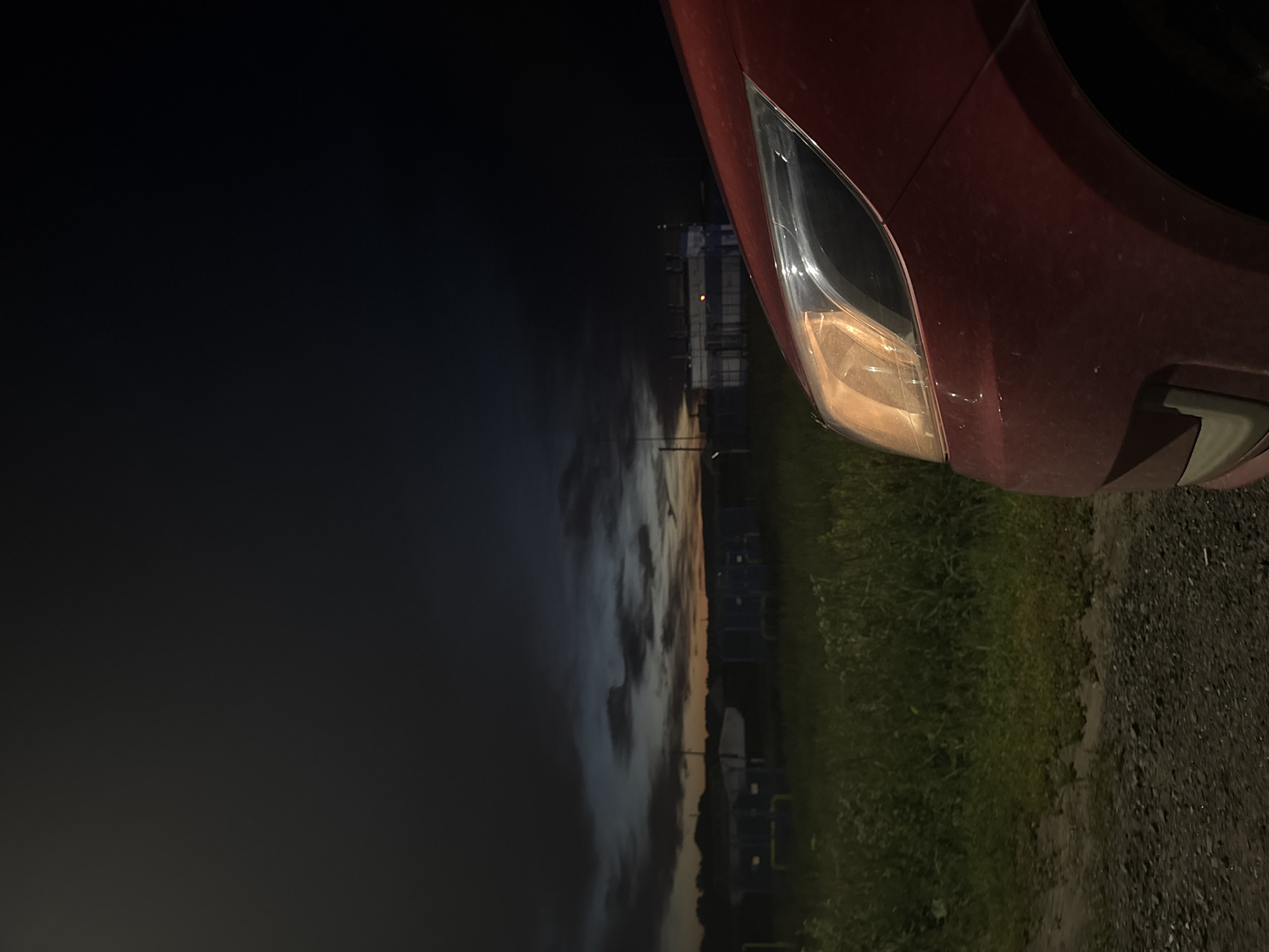 An exterior, low-angle, eye-level shot features the front right fender and headlight of a red car at night. The car's headlight is illuminated, casting a warm glow. The background consists of a dark, cloudy sky with hints of orange and blue near the horizon, suggesting either dawn or dusk. In the middle ground, there is a grassy field with some industrial-looking structures, possibly storage containers or small buildings, painted in blue. The ground in the foreground is a gravel road. The image is set in a rural or semi-industrial area. There are no people visible.