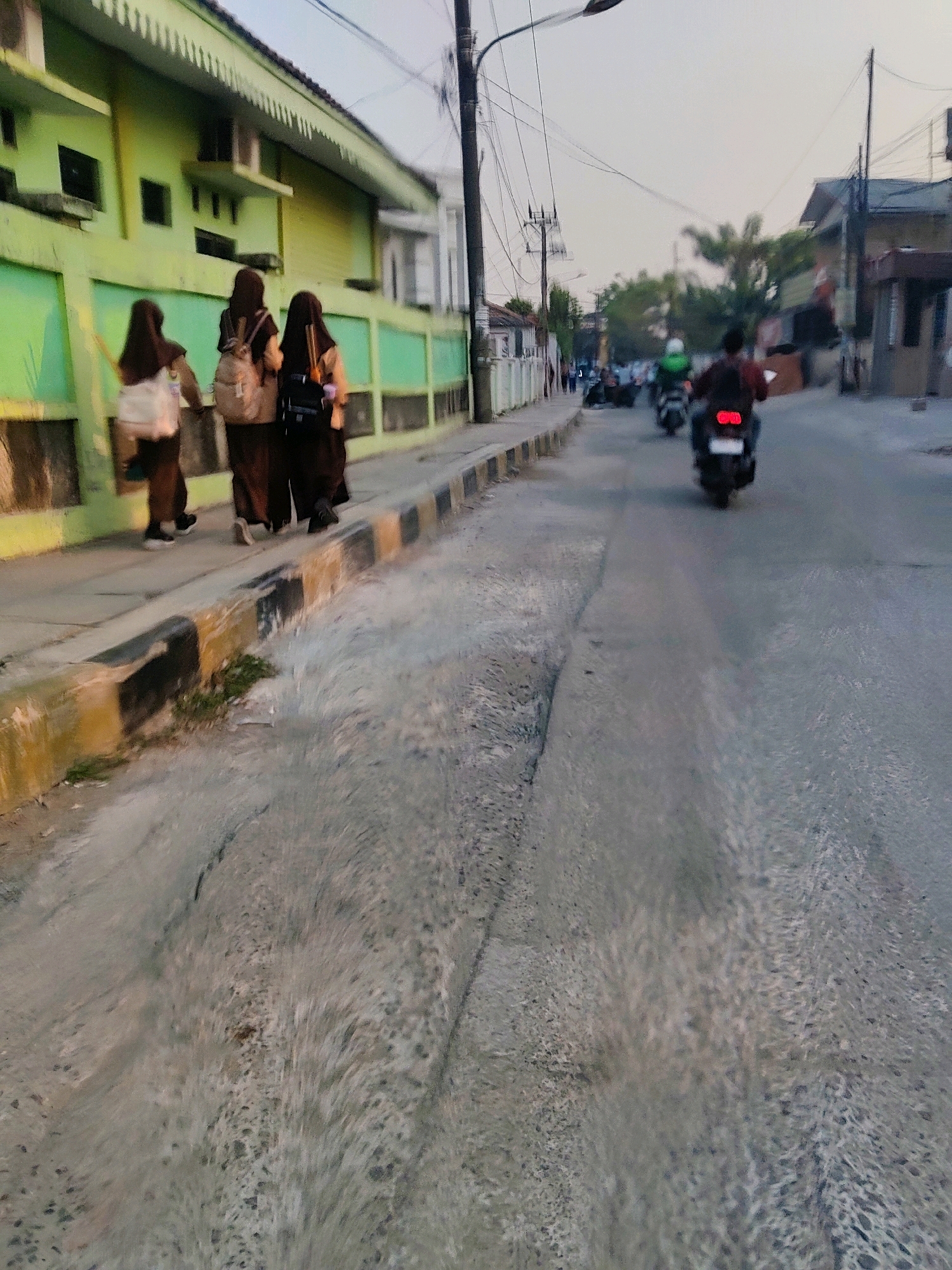 In Mekarmukti, Indonesia, three individuals are walking along a sidewalk adjacent to a bright green building, with a cracked asphalt road to their right. The individuals, likely students, are wearing hijabs and school uniforms, with two carrying backpacks. One person is wearing a white bag, another a tan backpack, and the third a black backpack.
The road is populated with several motorcycles, with one rider in the foreground clearly visible, their motorcycle's red taillight illuminated. The environment suggests late afternoon or early evening due to the soft, diffused light. The building to the left appears to be a school or similar institutional structure with visible windows and air conditioning units. The sidewalk is bordered by a curb with alternating black and yellow stripes. The road itself shows signs of wear and tear, with visible cracks and patches. Overhead, power lines and a street lamp are present, indicating an urban or semi-urban setting.