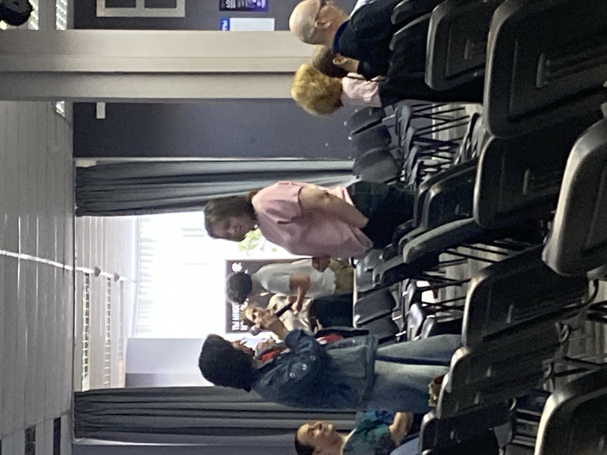 The image shows an indoor event, likely a lecture or presentation, taking place in Moscow, Russia. In the foreground, a woman with dark hair is holding a microphone and speaking to another woman in a pink shirt. Both women are standing. Behind them, other attendees are seated in rows of chairs. The room has grey curtains and a large window in the background, suggesting a bright, possibly daylight setting. The overall atmosphere appears casual and engaging.