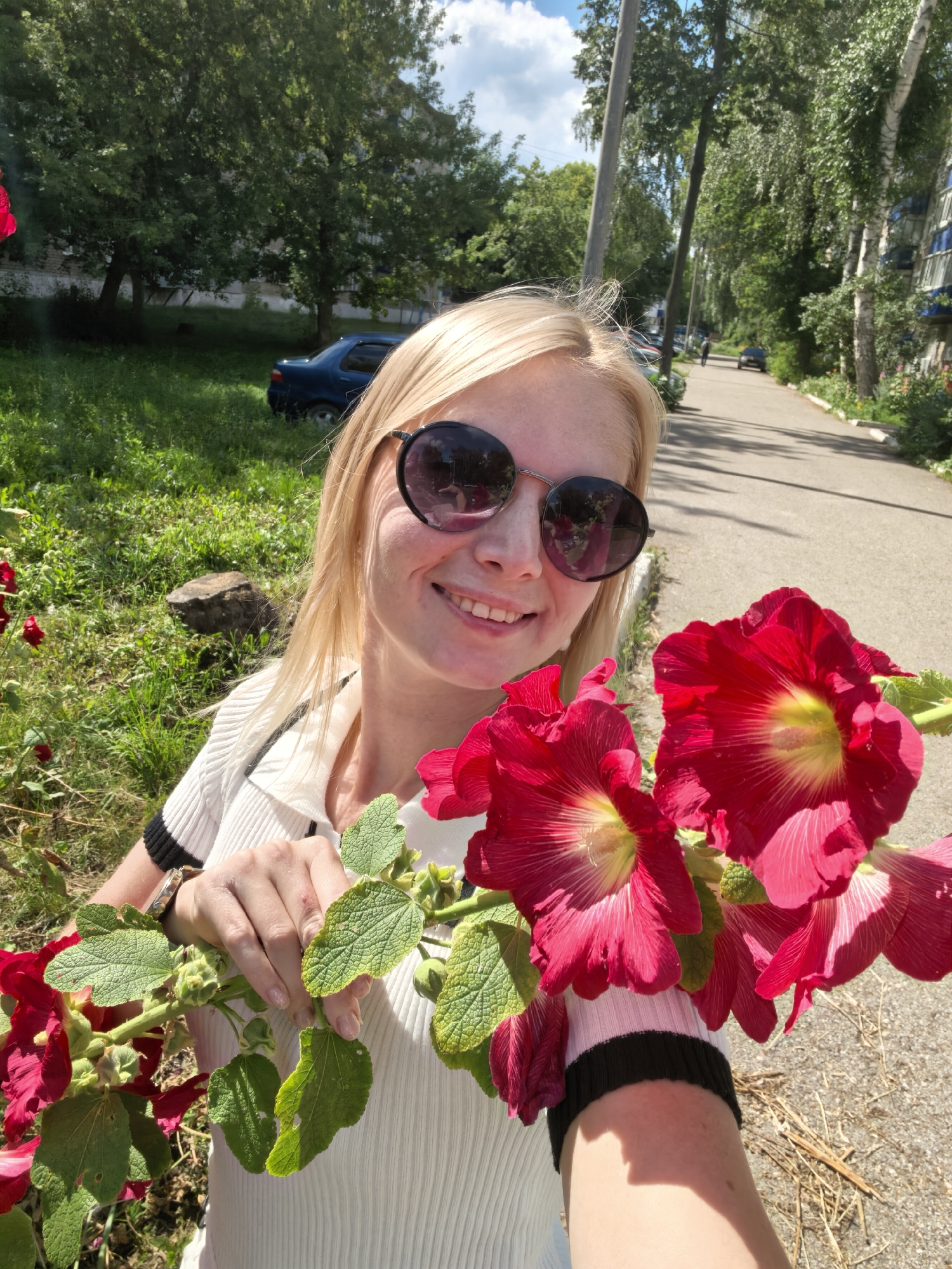 A blonde woman with sunglasses is taking a selfie outdoors in Sterlitamak, Russia. She is holding red flowers and wearing a white top with black trim on the sleeves. Behind her, a paved road curves to the right, lined with trees and a multi-story building. Green grass and a parked blue car are visible to the left. The sky is blue with some clouds.