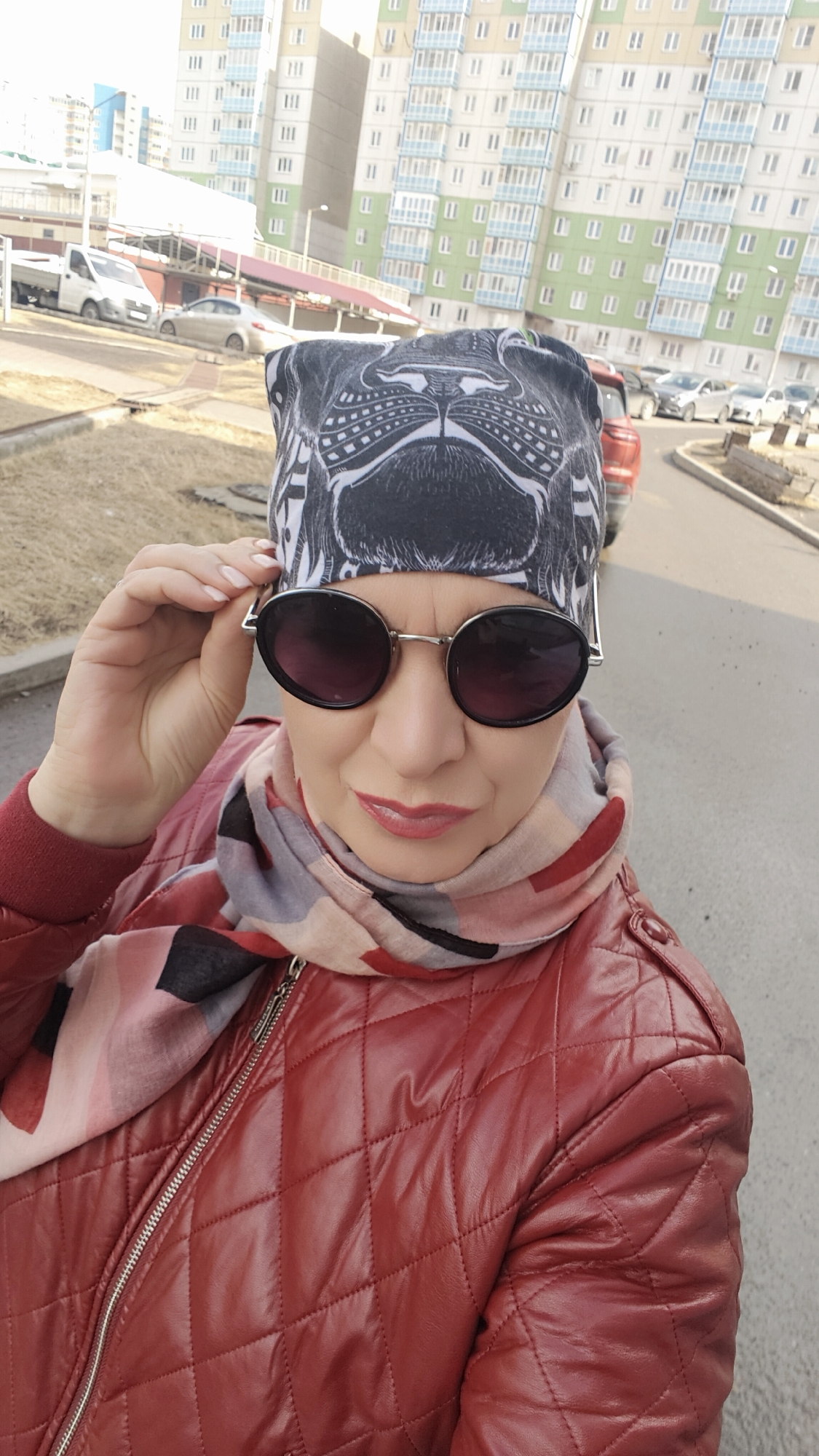 A woman wearing a red quilted jacket, a scarf, sunglasses, and a black-and-white patterned head covering is seen in an eye-level shot outdoors. She is adjusting her sunglasses with her left hand, and her gaze is directed towards the viewer. The woman has light skin, red lipstick, and appears to be middle-aged.
Behind the woman, the background consists of a cityscape. There are several buildings with light-colored facades and multiple windows. Cars are parked along the side of the road. The lighting indicates it is daytime.
The location appears to be an urban setting.