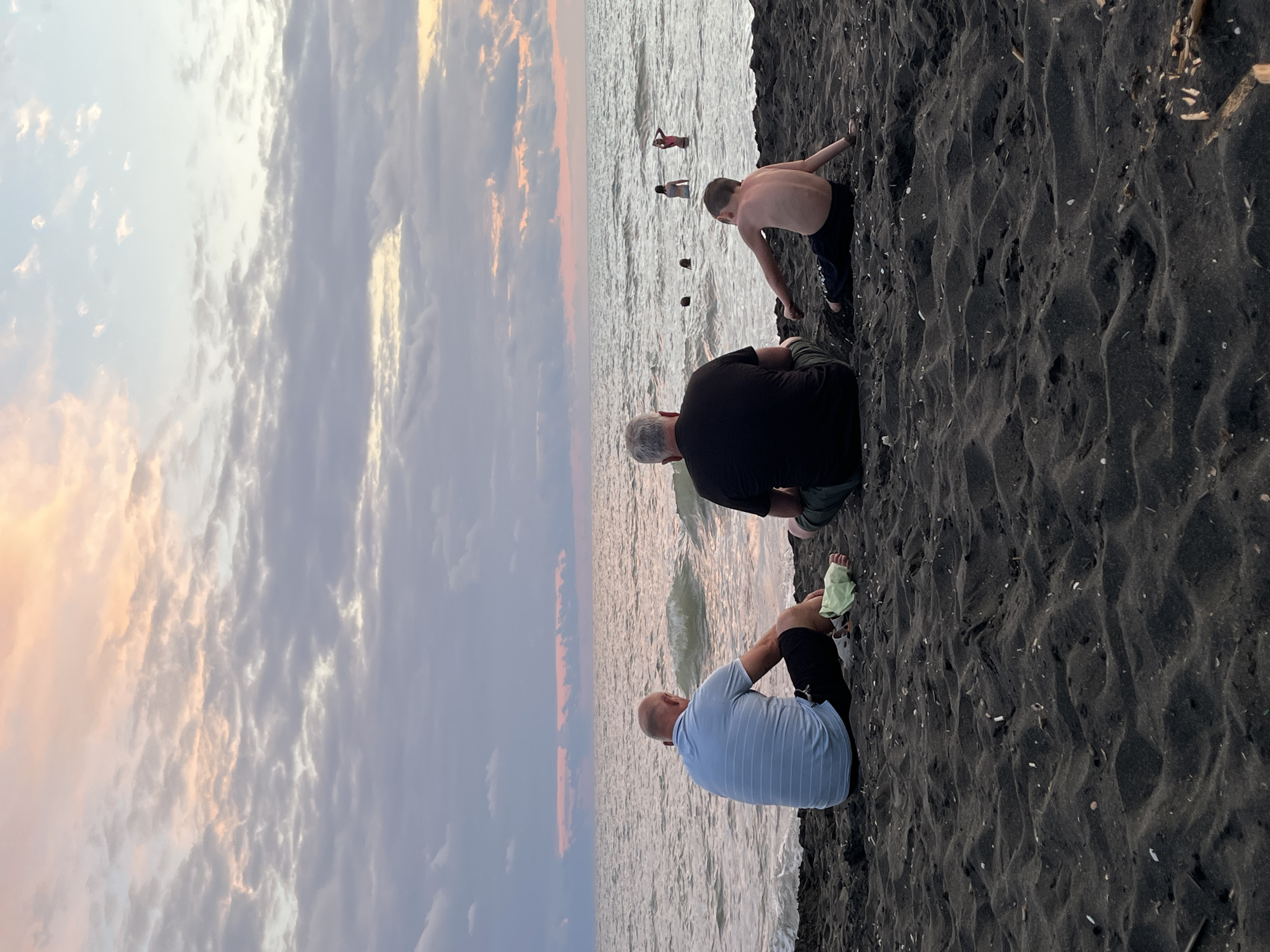 The image depicts a beach scene in Ureki, Georgia, during what appears to be sunset or sunrise. Three individuals are seated on the dark sand, facing the sea. On the left, a man wearing a light blue striped shirt and dark shorts is seated, with a pale green object on the sand near his feet. In the center, another man in a black t-shirt and green shorts is seated. To the right, a young boy in dark shorts is seated with his back to the camera, facing the water. In the sea, several other figures are visible, some appearing to be swimming or wading. The sky is filled with clouds tinged with pink and orange, reflecting on the water. The sand is dark and appears to be composed of fine particles.