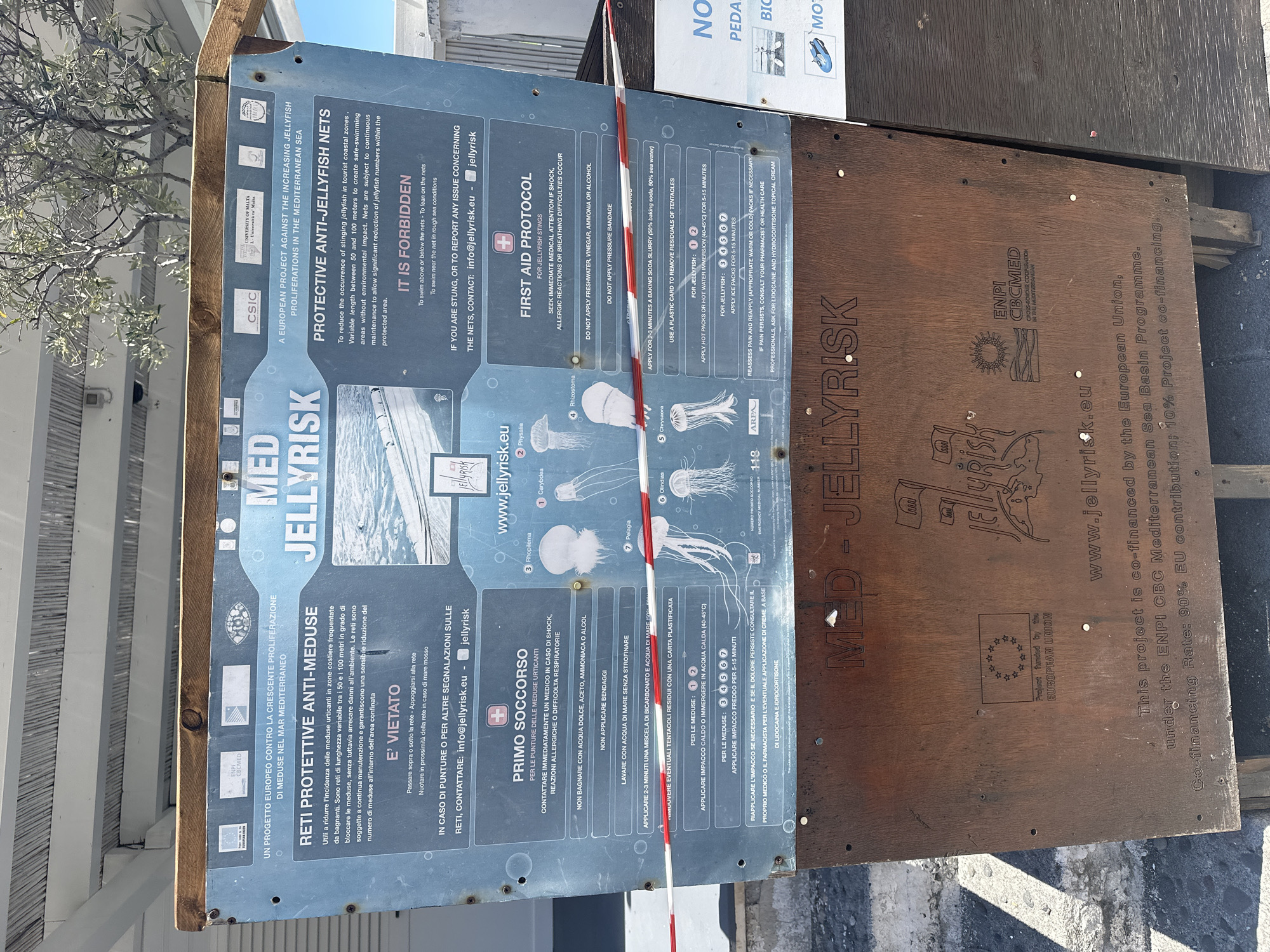 The image shows an information board about jellyfish risks, titled "MED JELLYRISK," displayed outdoors in what is presumed to be Lipari, Italy, based on the sign's content and context. The sign, written in both Italian and English, warns about jellyfish and provides first aid instructions for stings. It is divided into sections addressing protective anti-jellyfish nets and protocols for handling jellyfish stings. The sign also includes images of various jellyfish species for identification and contact information for reporting concerns. The board is affixed to a weathered wooden backing that has the "MED JELLYRISK" logo carved into it. The board has an information sign indicating that it is partly funded by the European Union. A red and white barrier tape is also visible, possibly to prevent access to the sign.