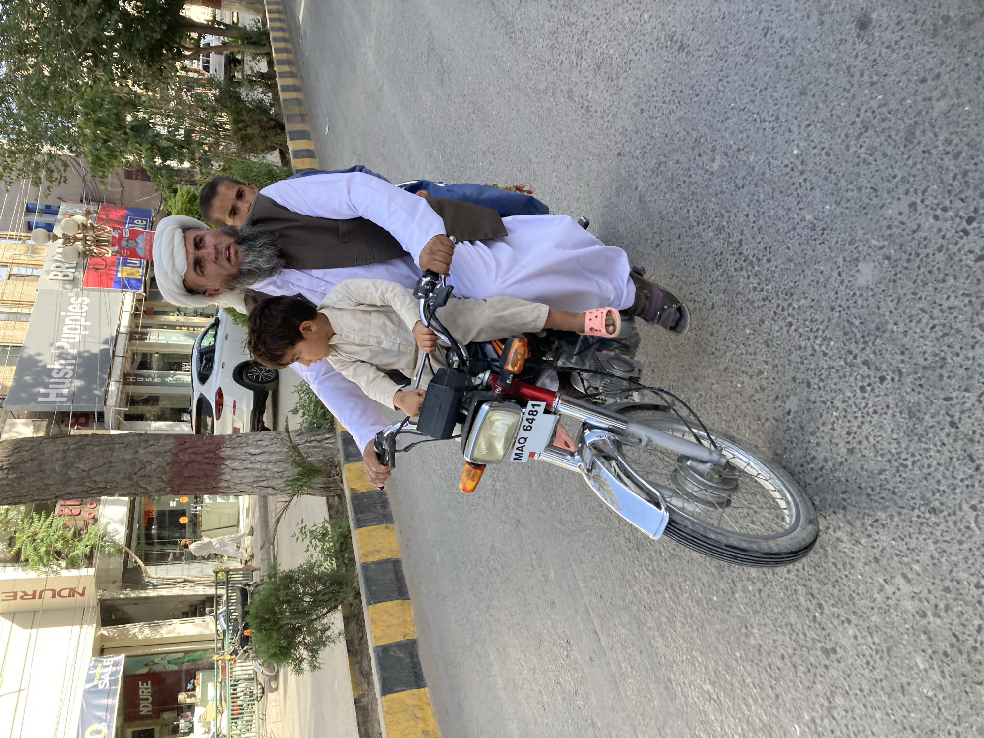 In Quetta City Tehsil, Pakistan, an adult male with a white turban, long grey beard, white clothing, and a brown vest operates a silver motorbike on a paved street. Two children accompany him, one sitting in front of him and holding the handlebars, and another sitting behind him. Buildings and a tree line the side of the street. The motorbike's license plate reads "MAQ 6481".