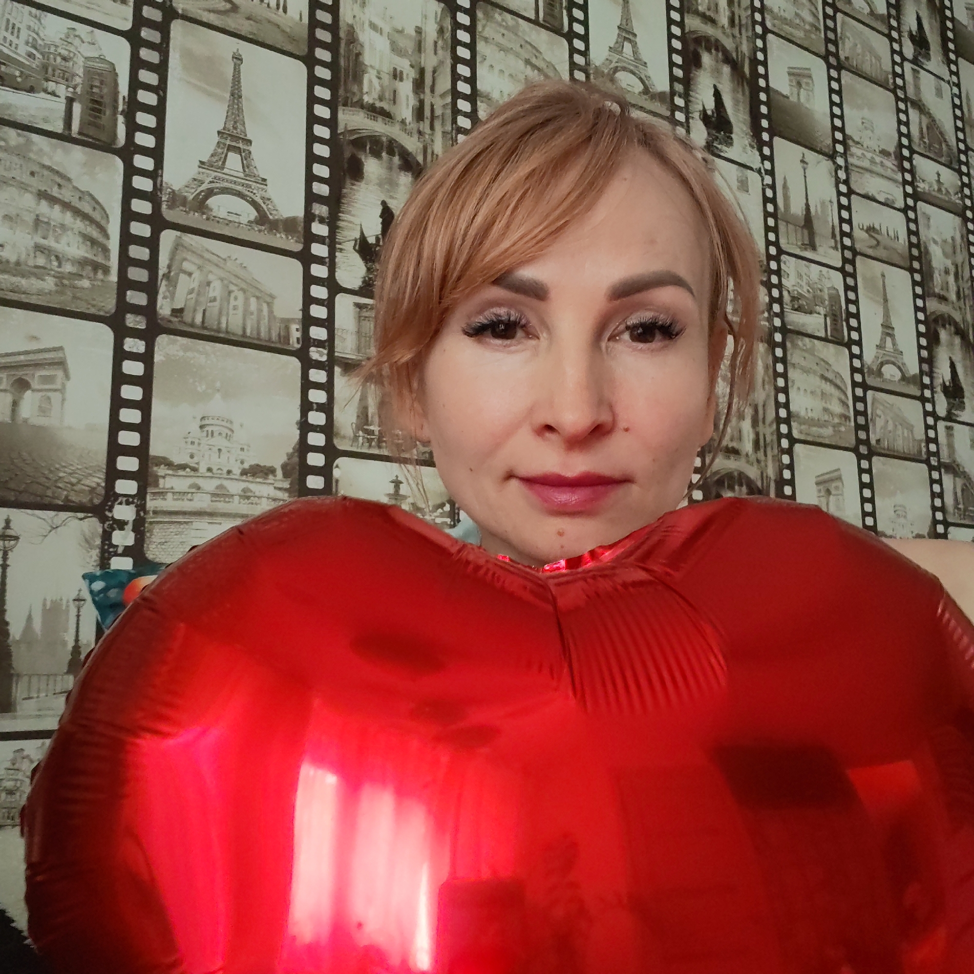 A woman is centered, holding a large red heart-shaped balloon close to her chest. She has fair skin, light brown hair pulled back, and is wearing makeup with visible eyelashes and lipstick. Her expression is neutral.
The background is a wall covered with wallpaper that features black and white images of various landmarks and buildings framed as film strips, including the Eiffel Tower and the Colosseum.
The location is Chaikovsky, Russia.