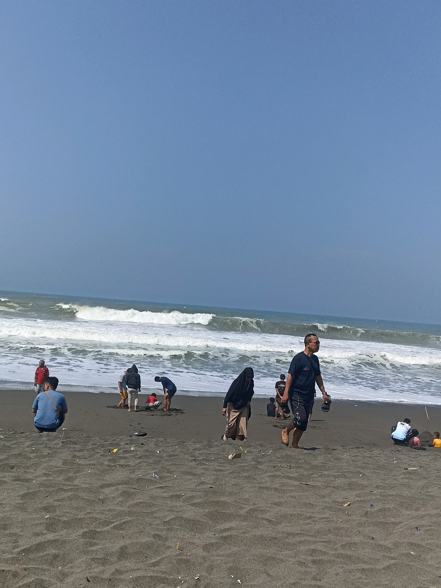 The image depicts a sandy beach in Kenoyajaya, Indonesia. The sky is a clear, pale blue. Waves are crashing onto the shore, creating white foam. Several people are present on the beach. In the foreground, a person wearing a blue shirt and shorts is crouched down, facing away from the camera. Further back, near the water's edge, a group of individuals are visible. One person in a black head covering and a long skirt is walking. A man in a dark t-shirt and shorts is walking towards the right side of the frame, holding a pair of flip-flops. Another person in a dark t-shirt and light-colored pants is also in this area. To the right, two people are kneeling on the sand, one wearing a white and blue top, and another possibly a child. A small child in a yellow shirt is sitting nearby. The sand is dark and appears somewhat wet near the water. Scattered across the sand are small pieces of debris, including plastic.