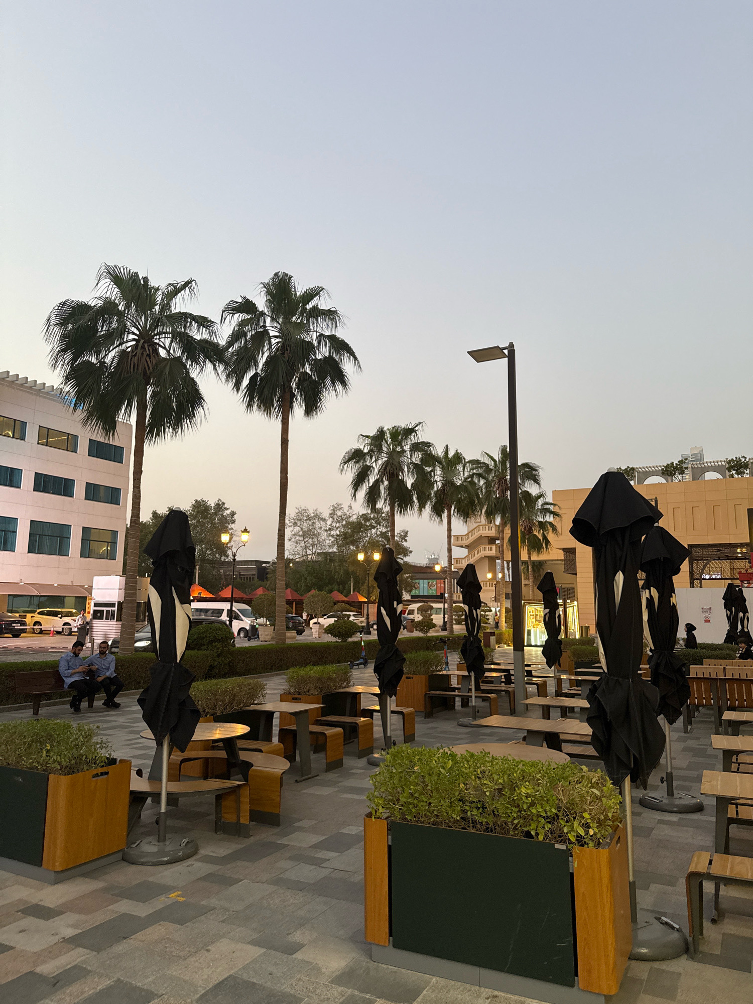 The image shows an outdoor dining area with several tables and chairs, many of which are covered by closed black umbrellas. The area is paved with patterned tiles and bordered by planters filled with greenery. In the background, there is a modern building with glass windows, palm trees, and a street lamp. The sky is a pale, hazy blue, suggesting either early morning or late evening. A few people are visible: two men are sitting on a bench on the left, and other figures, some partially obscured, are scattered throughout the scene, possibly patrons or staff. Cars and vans are parked in the distance. The overall impression is of a public space, likely a commercial or restaurant district, given the presence of outdoor seating and the style of the buildings. This scene is characteristic of an urban environment in Dubai, United Arab Emirates, where such outdoor dining areas are common and palm trees are a prevalent feature of the landscaping.