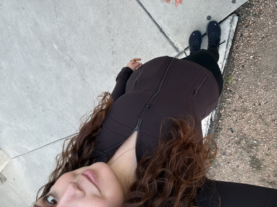 A woman stands on a sidewalk near a curb. She wears a fitted brown jacket with a zipper down the front, black leggings, and black boots. Her curly brown hair cascades around her face and down her chest. The sidewalk is concrete, and the curb is concrete and dirty. The photograph is taken from a high angle looking down at the woman.