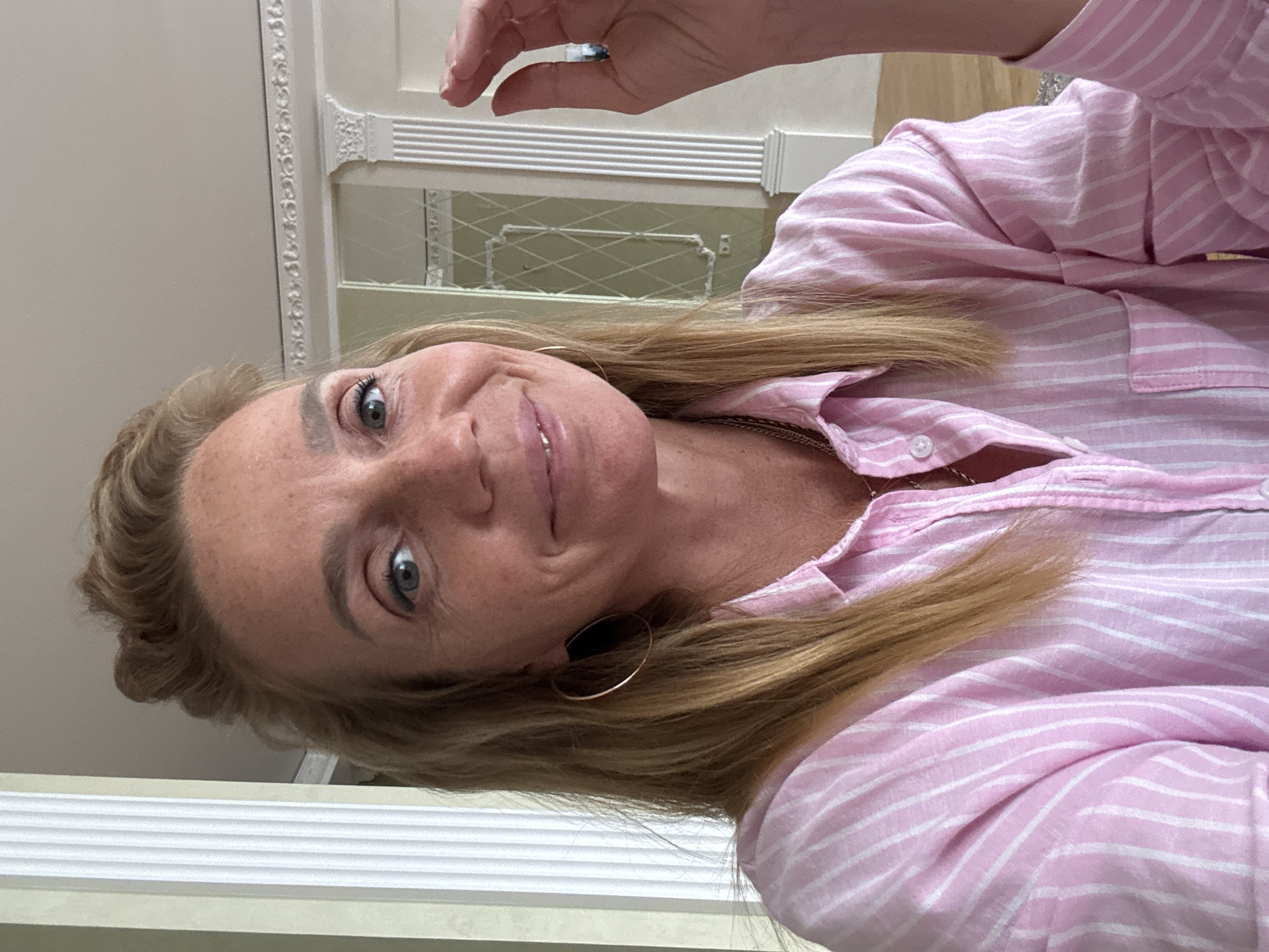 A middle-aged Caucasian woman with blonde hair pulled back is looking at the camera, smiling. She is wearing a pink and white striped button-down shirt and gold hoop earrings. Her right hand is raised, with her index finger and thumb close together, as if she is indicating a small size. She is indoors, with ornate white molding and pillars visible in the background, suggesting an interior in an older, possibly grand, building. The lighting is soft and appears to be natural. There is no indication of the city of Sinyavino, Russia, in the image.