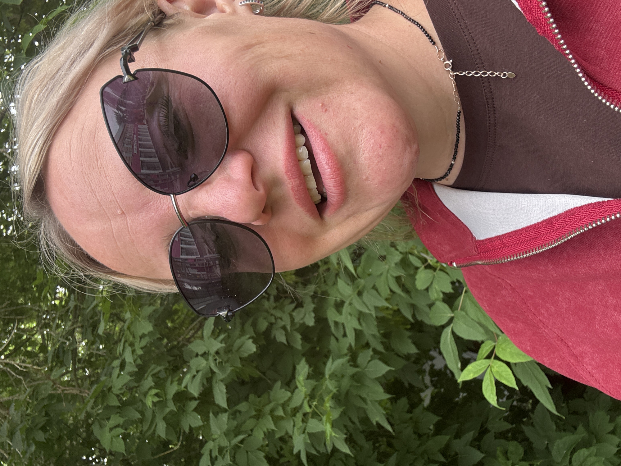 The image is a close-up selfie of a woman with blonde hair and fair skin, taken outdoors against a backdrop of lush green foliage. She is wearing large, dark-lensed sunglasses with a distinctive angular frame, through which reflections of a building with multiple windows are visible. Her mouth is slightly open, revealing her teeth and pink lipstick. She has a delicate necklace made of small black beads and a silver chain around her neck. She is also wearing a red zippered jacket over a brown shirt and a small silver hoop earring in her left ear. The lighting suggests it is daytime. The location is the city of Chastyye, Russia.