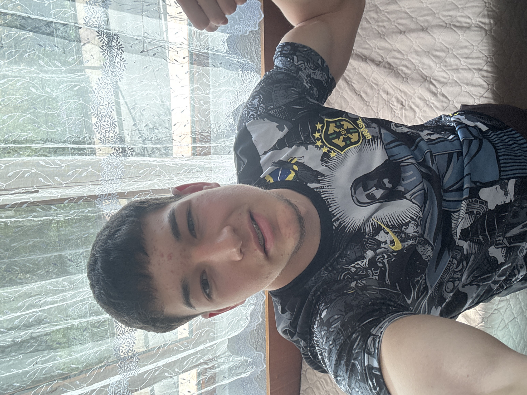 This media file is a selfie of a young man, taken indoors in Zlatograd, Bulgaria, during daylight hours. The man is positioned on his side, looking directly at the camera with a slight smile. He has short, dark hair, light facial stubble, and is wearing visible braces on his upper teeth. A few red blemishes are noticeable on his forehead and left cheek.
He is wearing a multi-colored t-shirt, predominantly black, grey, and yellow, with a graphic design. The design prominently features a crest with five stars, the text "CBF BRASIL" and "BRASIL," which are hallmarks of the Brazilian Football Confederation. Various stylized figures and patterns are also integrated into the shirt's print. His right arm is bent, and his hand is partially visible at the top of the frame, suggesting he is holding the camera to take the photo.
The background features a window covered by a sheer white curtain with an intricate embroidered leaf pattern. Through the translucent curtain, hints of outdoor greenery, possibly trees, and window frames are visible, indicating an exterior view. To the right, a textured, light-colored quilted material, resembling a bedspread or blanket, occupies part of the frame, suggesting the man might be on a bed or sofa. The scene is brightly lit by natural light streaming through the window, confirming it is daytime.