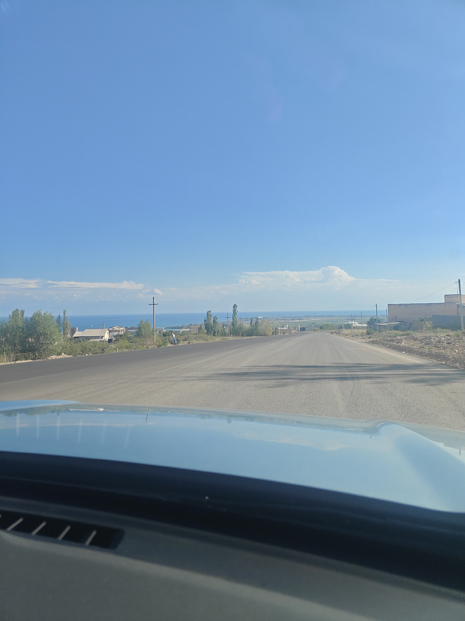 The image captures a view from a vehicle driving on a paved road towards a body of water, likely Lake Issyk-Kul, near Cholpon-Ata, Kyrgyzstan. The sky is a clear, bright blue with a few wispy clouds near the horizon. The road stretches into the distance, bordered by a dusty shoulder. On the left side of the road, there are trees and some buildings, suggesting a settlement. On the right, a sandy embankment with scattered construction materials is visible, along with a brick building. Power poles are present along the roadside. The reflection of the sky and surrounding environment is visible on the vehicle's dashboard and hood.