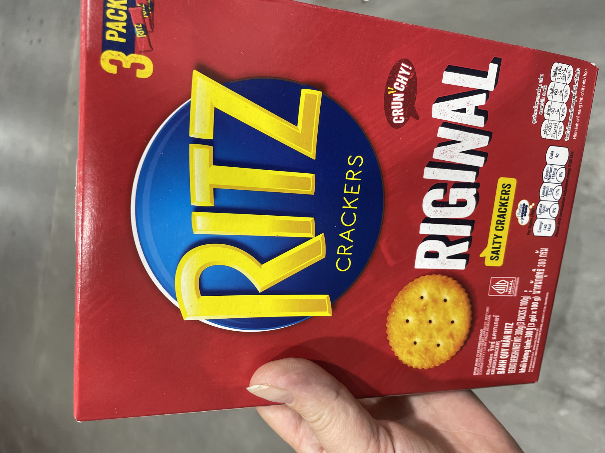A close-up, top-down, slightly angled exterior shot captures the front of a red Ritz Crackers box held by a person's left hand and fingers. The box features the iconic Ritz logo in large, yellow, uppercase letters against a blue circle, with "CRACKERS" written in yellow underneath. The box is also marked with "3 PACK" in yellow text. A single, round, golden-brown cracker with visible perforations is depicted on the lower left side of the box. Text in various languages, including English, Thai, and Vietnamese, is visible on the box, indicating product details and nutritional information. A small, red, speech bubble graphic with "CRUN'CHY!" written in white text is positioned on the right side. The background is out of focus, showing a greyish, textured surface, likely a table or counter. The presence of the Ritz crackers box strongly suggests a retail or home environment where the product is displayed or stored. The scene is well-lit, with no visible environmental damage or hazardous conditions. The hand is clean and appears to be that of an adult. The overall impression is that of a typical consumer product presentation.