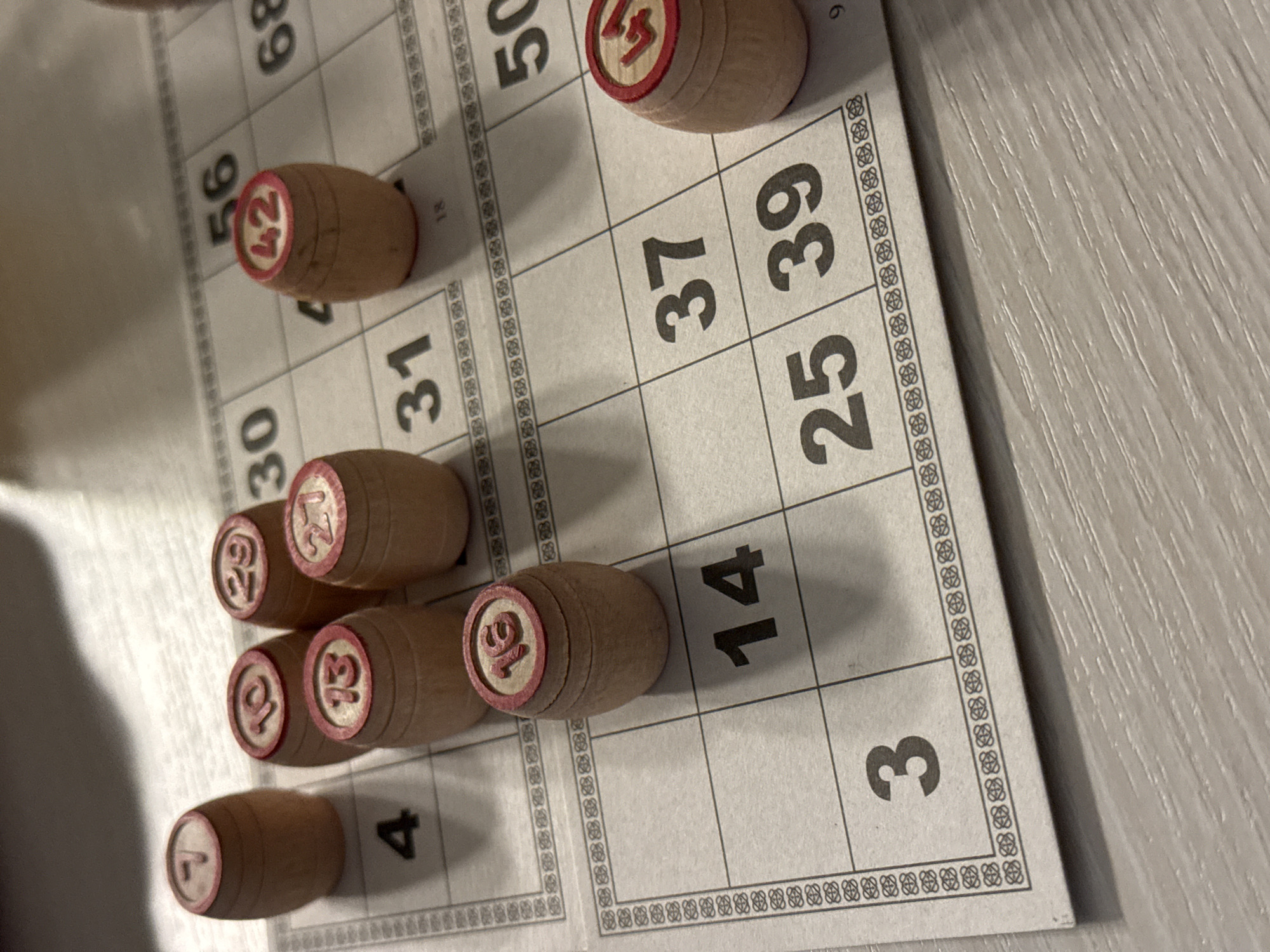 A close-up shot depicts a bingo game in progress. The game board is visible, with numbers ranging from 3 to 68. Several wooden barrels with numbers printed on them are placed on the board, marking the called numbers. The barrels display the numbers 1, 12, 13, 16, 27, 29, 42, and 53. The bingo card displays the numbers 3, 4, 14, 25, 30, 31, 37, 39, 50, 56, and 68. The photo is taken at an angle, showing the bingo game on a light-colored table, likely indoors. There is no indication of the city of Chaikovsky, Russia.