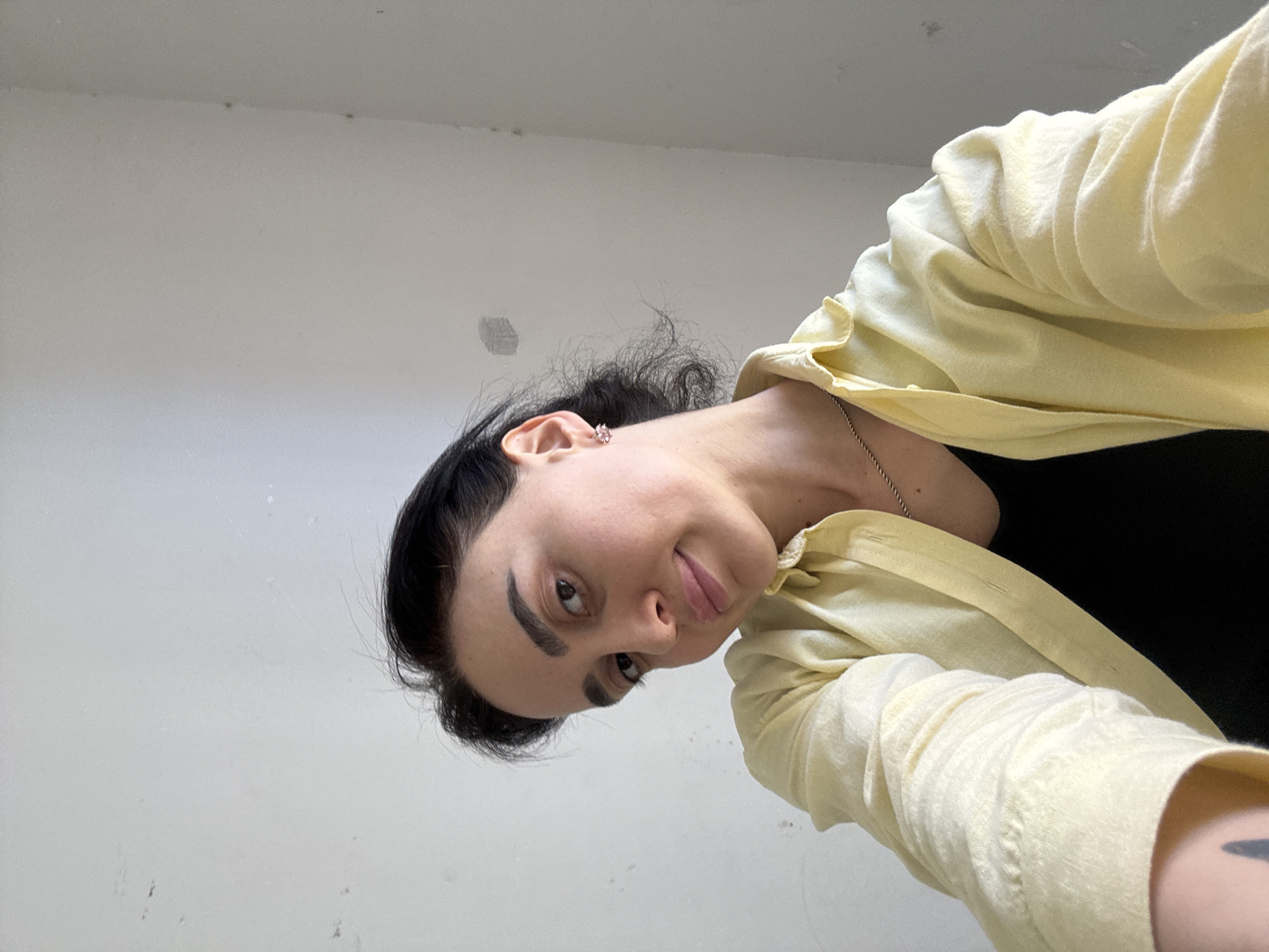 A selfie taken indoors shows a person with dark hair, wearing a yellow collared shirt over a black top, and a silver necklace. The person has dark eyebrows, fair skin, and is wearing a small earring. The background consists of a white wall with some slight imperfections. The location cannot be determined from the image.