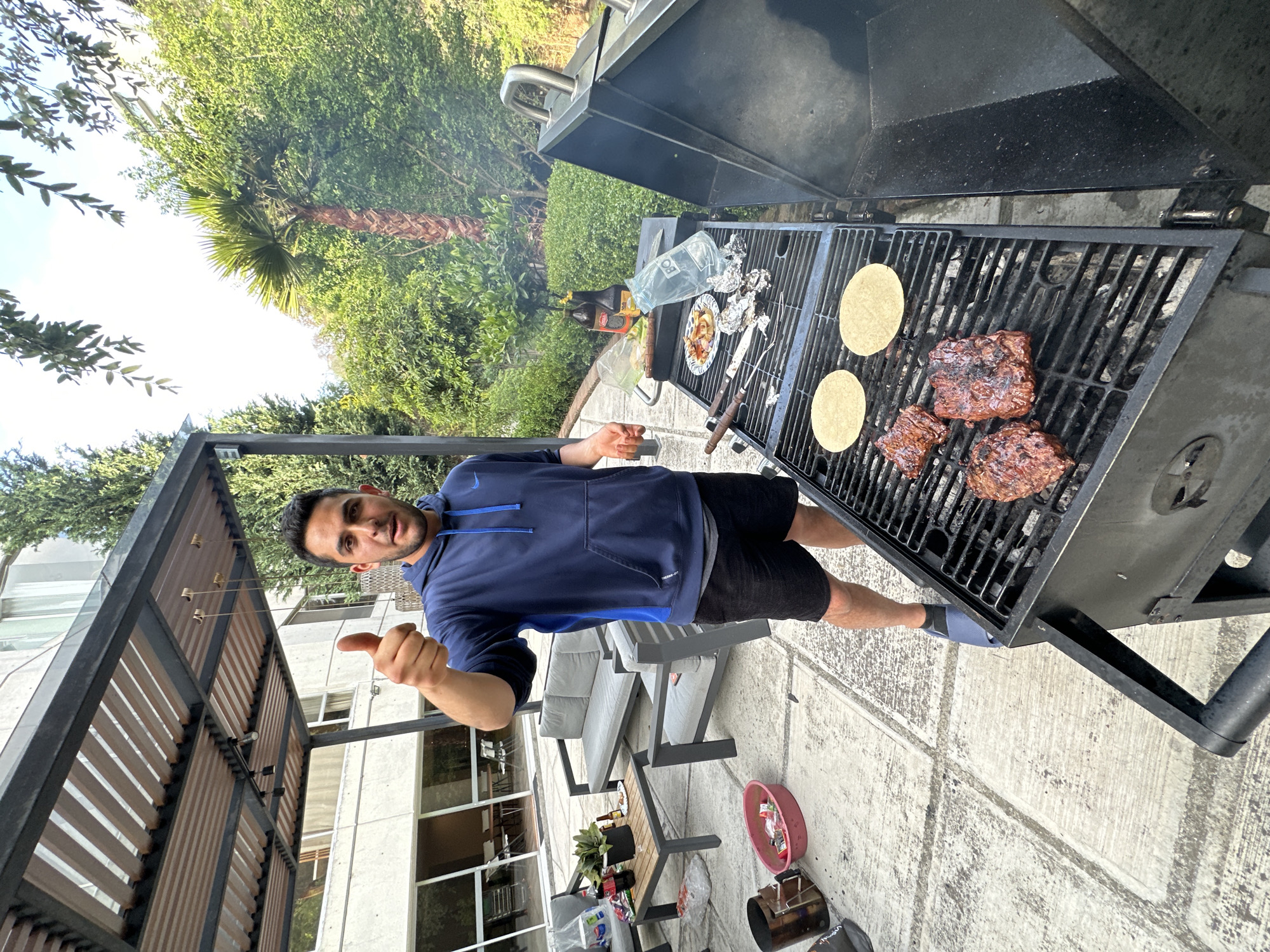 A man stands outdoors, giving a thumbs-up gesture near a barbecue grill in Huixquilucan de Degollado, Mexico. The grill contains two tortillas and several pieces of meat. Other items are grilling on a second level of the grill including some foil wrapped packages, a plate of food, and various sauces. The setting includes a patio with a pergola, outdoor furniture, and a view of greenery.