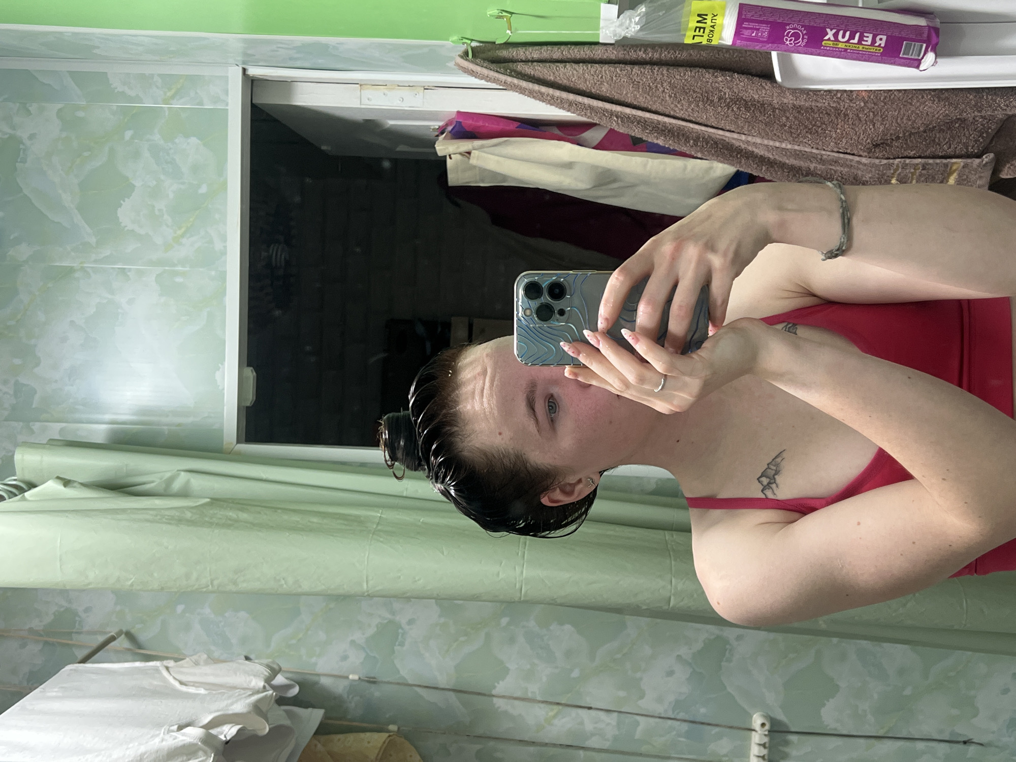 A woman is taking a selfie with her phone in a bathroom. The bathroom has green and white marble patterned walls. There is a mirror behind her. Clothes are hanging on a rack on the wall on the bottom left corner. A brown towel and various items are on a shelf above her head. There are no other indicators that can confirm the location as the city of Chaikovsky, Russia.