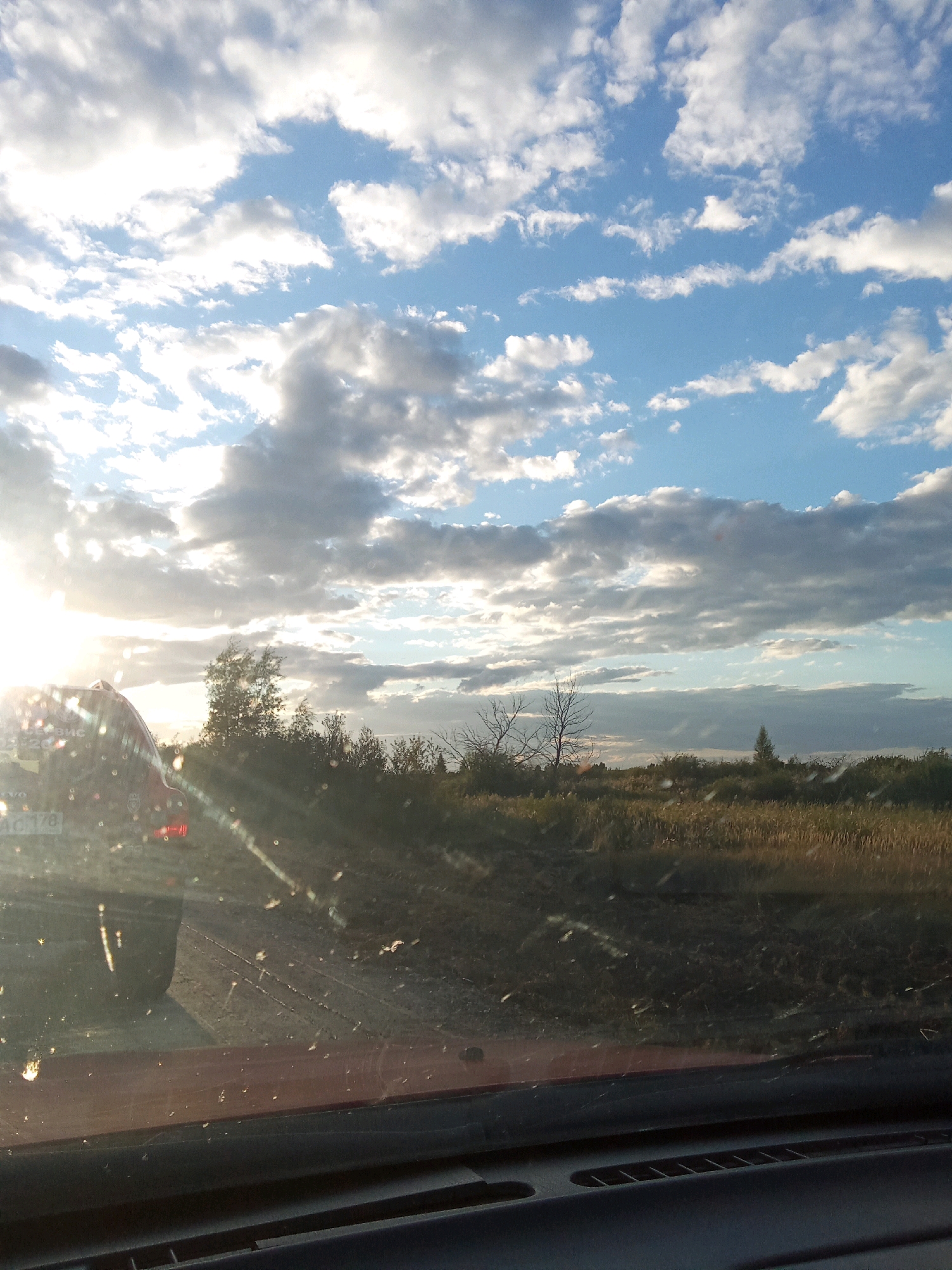 The image depicts a sun-drenched landscape with a partially obscured view of a vehicle's interior in the foreground. The sky is a vibrant blue, dotted with dramatic, sunlit clouds. A bright sun casts long rays, suggesting it is either early morning or late afternoon. The scene appears to be an open, rural area, possibly on the outskirts of Бәйтерек ауылдық округі, Kazakhstan, given the presence of dry, tall grass and scattered trees and bushes. A red car is visible on the left, with its rear and side partially in frame. The vehicle's windshield is notably dirty, with numerous small spots and streaks. The overall impression is one of a journey through a natural, possibly arid, environment under a dynamic sky.