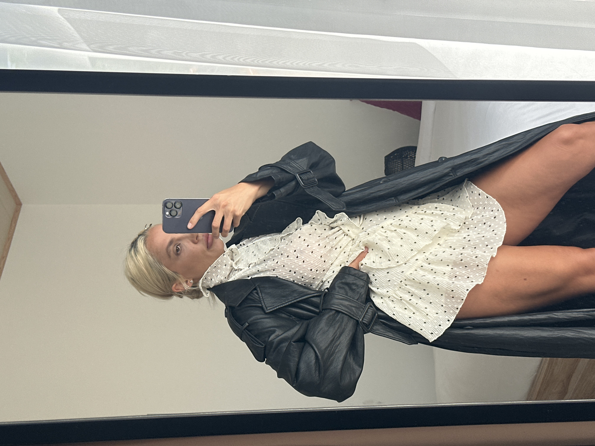 A woman is taking a mirror selfie in Kuta Selatan, Indonesia. She is wearing a black leather trench coat over a white dress with black polka dots and ruffles. The dress appears to be short. She is holding a dark-colored smartphone in her right hand. Her left hand is on her hip. The background shows a room with a bed, a white curtain, and a window. The mirror she is using is large and framed in black. The lighting is bright, suggesting it is daytime.