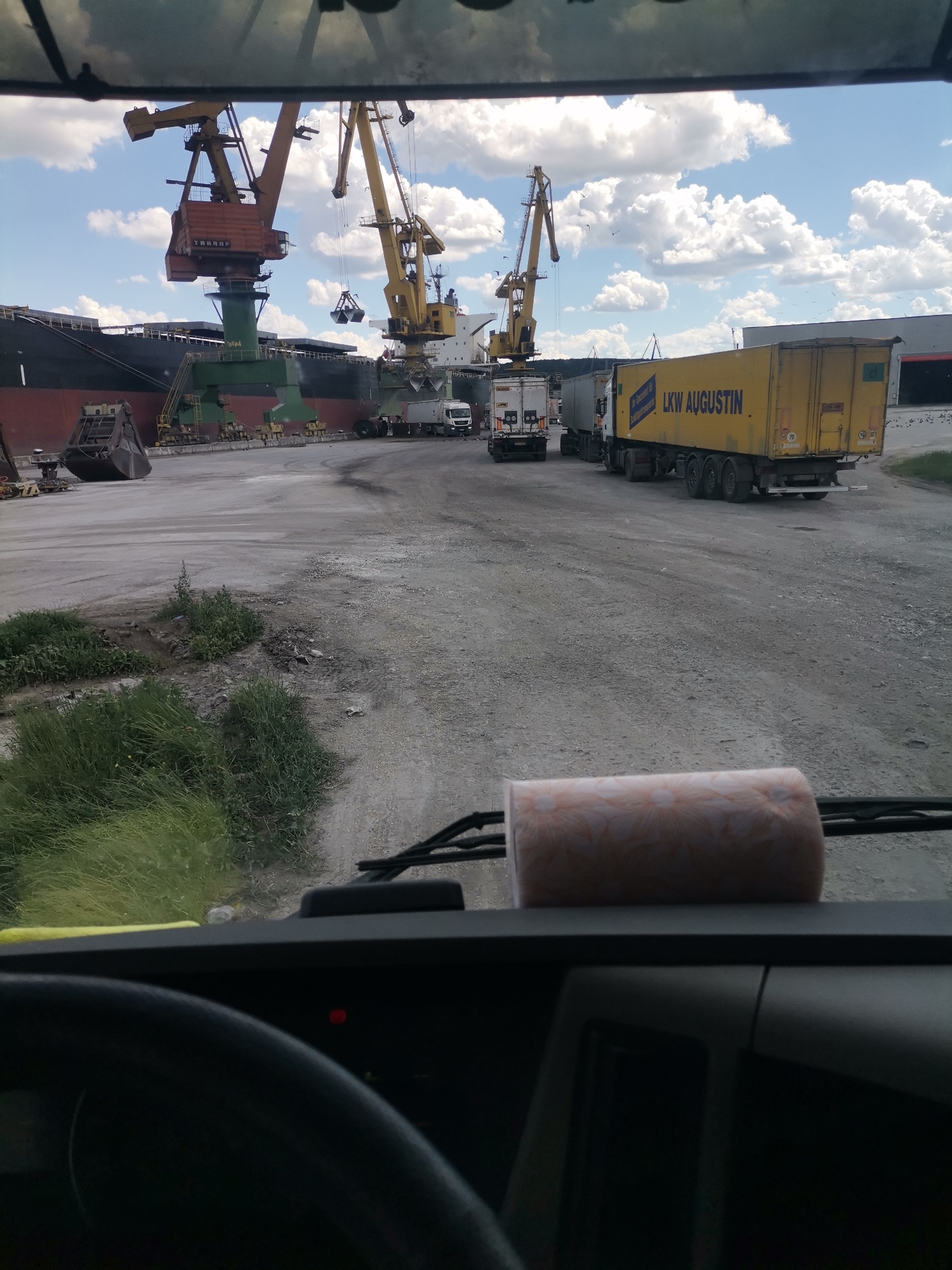 The image shows a view from inside a vehicle, looking out at a port or industrial area. In the background, large cranes are positioned next to a docked ship. Several trucks are visible, including a yellow truck with "LKW AUGUSTIN" written on its side. The ground is a dusty, unpaved surface, and there is some vegetation on the left side of the image. The sky is blue with white clouds. The scene appears to be in a working industrial environment, likely a port, where cargo is being loaded or unloaded from a ship using cranes and transported by trucks. The location is identified as Ezerovo, Bulgaria, which is consistent with a port or industrial area near a body of water.