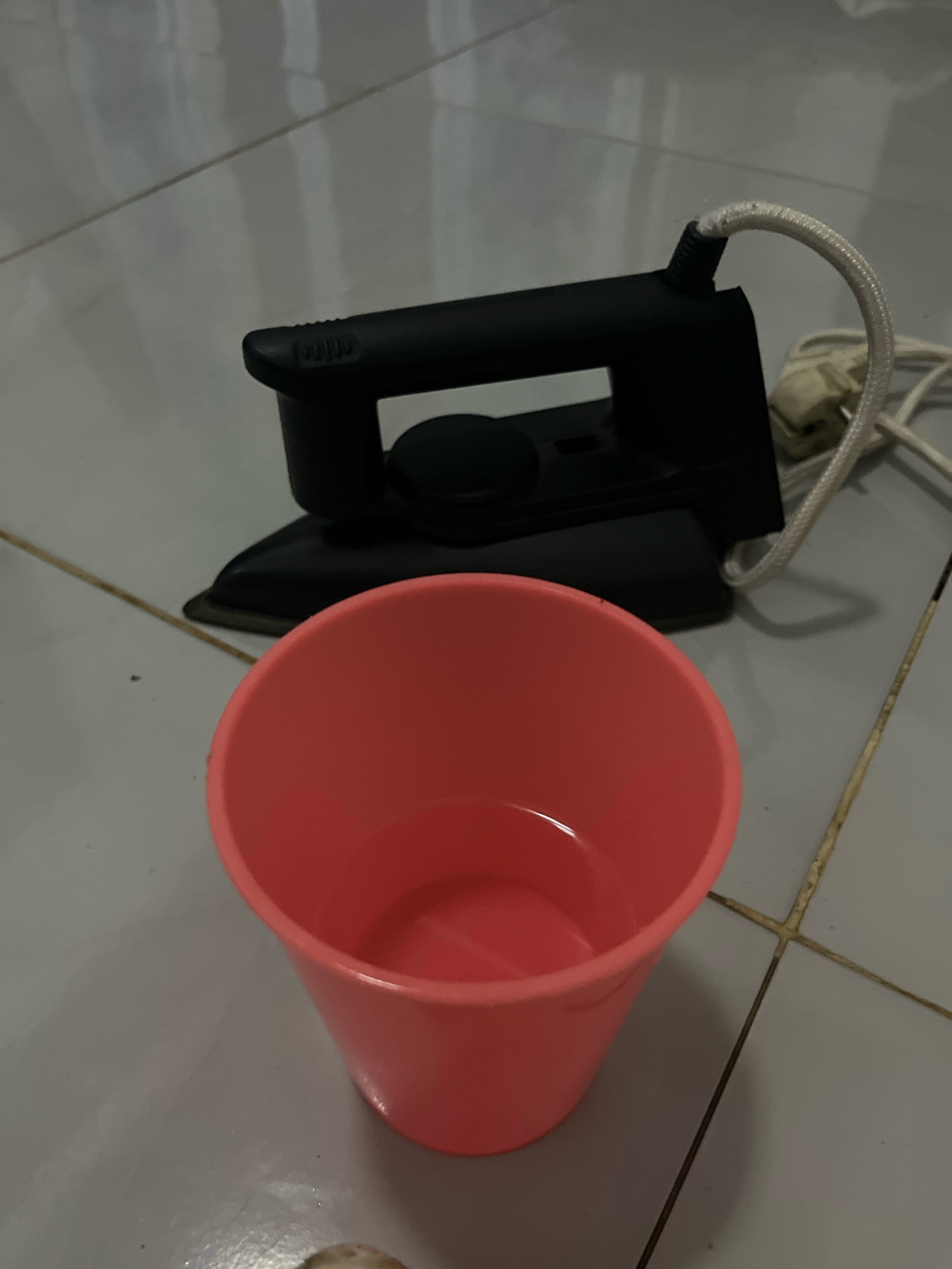 A black steam iron rests on a tiled floor, its power cord trailing. In the foreground, a bright pink plastic cup filled with water stands beside the iron. The scene appears to be indoors, possibly in a home or a laundry area, given the presence of the iron and the tiled flooring common in Indonesian households. The image does not provide specific indicators to pinpoint its exact location within Kebumen, Indonesia, beyond the general context of a domestic setting.