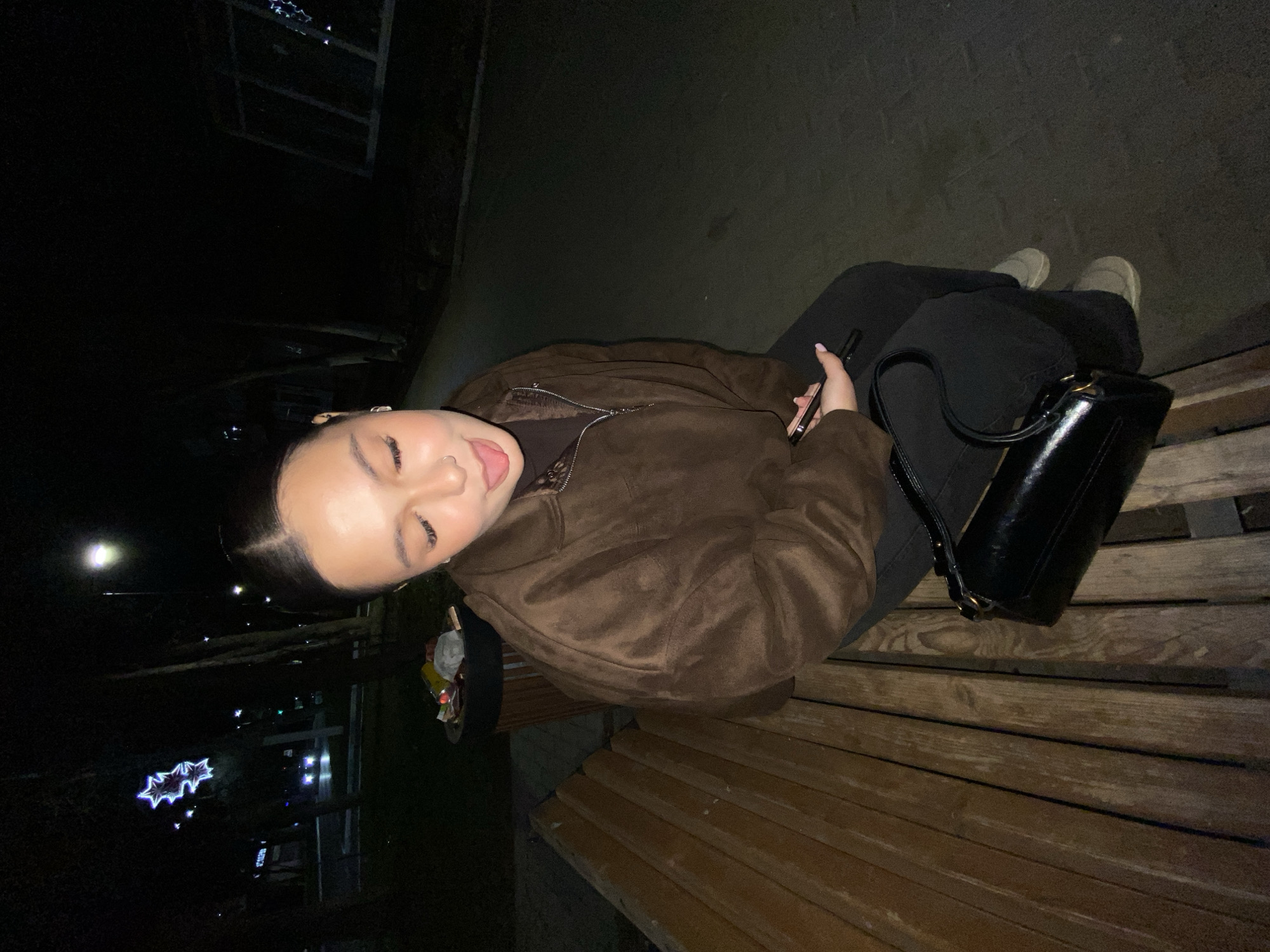 A young woman sits on a wooden bench, likely in a park. She is wearing a brown jacket and black pants. A black purse sits next to her. She is holding a phone in her right hand. Her tongue is sticking out. The location is Chaikovsky, Russia. The setting appears to be at night. There is a tree or structure behind her with decorative lights.