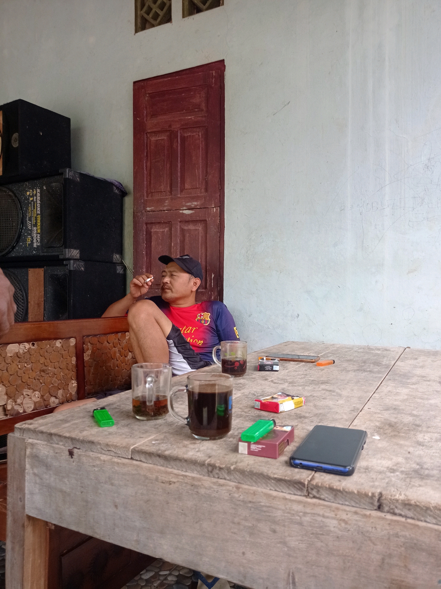 A man is smoking a cigarette and sitting on a bench next to a table laden with drinks, cigarettes, and a phone. The scene is set outdoors in Kalialang, Indonesia, with a light-colored wall and a red wooden door in the background. The man wears a blue and red t-shirt and a black baseball cap. Two mugs of tea and coffee sit on the table, alongside two packs of cigarettes, a lighter, and a mobile phone. A stack of black speakers is visible to the left of the man. The table is made of rough wood, and the bench has intricately carved wooden panels. The wall is plain and shows some scuff marks.