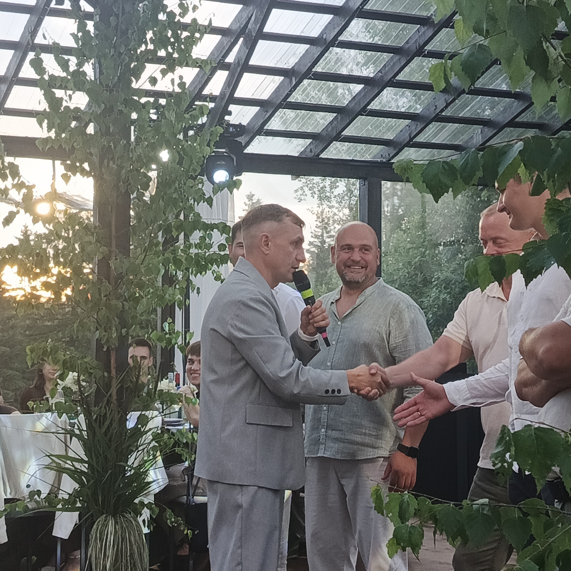 The image depicts an outdoor event, possibly a wedding reception or a corporate gathering, taking place in the city of Svetloye, Russia. The scene is set under a wooden pergola with a transparent roof, suggesting it's an open-air venue. Several men are visible, some engaged in conversation and handshakes, indicating social interaction. One man in a grey suit is holding a microphone and shaking hands with another man wearing a light-colored shirt. In the background, more people are seated at tables, and greenery is abundant, with trees and plants framing the scene. The lighting suggests it is late afternoon or early evening, with the sun casting a warm glow through the foliage.