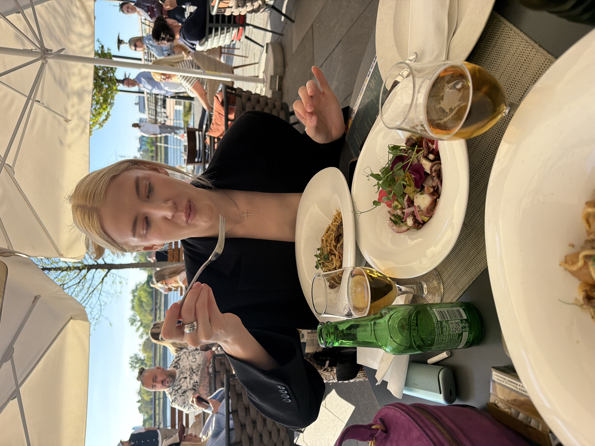 A woman with blonde hair is sitting at an outdoor table, eating and looking at the camera. She is holding a fork in her right hand. The table is set with several plates of food, including a salad with octopus, pasta and another plate of food that is only partially visible. There are also two glasses of beer and a green bottle on the table. The table is on a patio, and in the background, other people are seated at tables along the Sava river at Beton Hala, Belgrade, Serbia.
