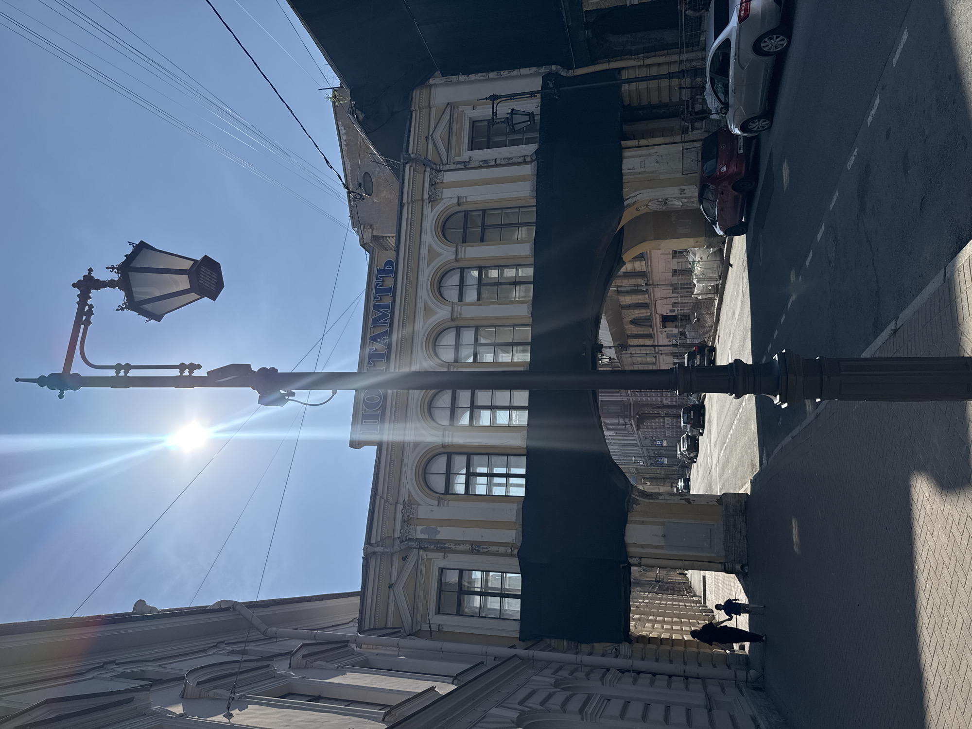 The image captures a street scene on a sunny day. In the foreground, a lamppost with an ornate design stands tall, partially obscuring a bright sun with visible lens flare. The lamppost's light fixture is angled, and its pole extends upwards, disappearing out of frame.
Behind the lamppost, an old building with classical architectural elements is visible. It features arched windows with multiple panes and decorative yellow and white trim. A large black tarp or banner is draped over a section of the building, creating a stark contrast against the sunny sky and the building's facade. The tarp appears to cover an archway or a passage.
To the right of the building, a paved street stretches into the distance. Several cars are parked along the side of the street, some with their license plates visible. The street is marked with white lines. The pavement on the sidewalk is made of bricks.
In the lower right portion of the image, two figures are silhouetted against the bright pavement. One is a larger figure, and the other, a smaller figure, appears to be walking alongside or holding hands with the larger one. They are walking away from the camera, casting long shadows.
The sky is clear blue, indicating a sunny day, likely in the morning or afternoon due to the sun's position and the strong shadows. The presence of overhead wires suggests an urban environment. Some text is visible vertically on the building, appearing to be in Cyrillic script. Part of the word "ТАМПЬ" is legible. The overall impression is of a historic urban setting bathed in bright sunlight.