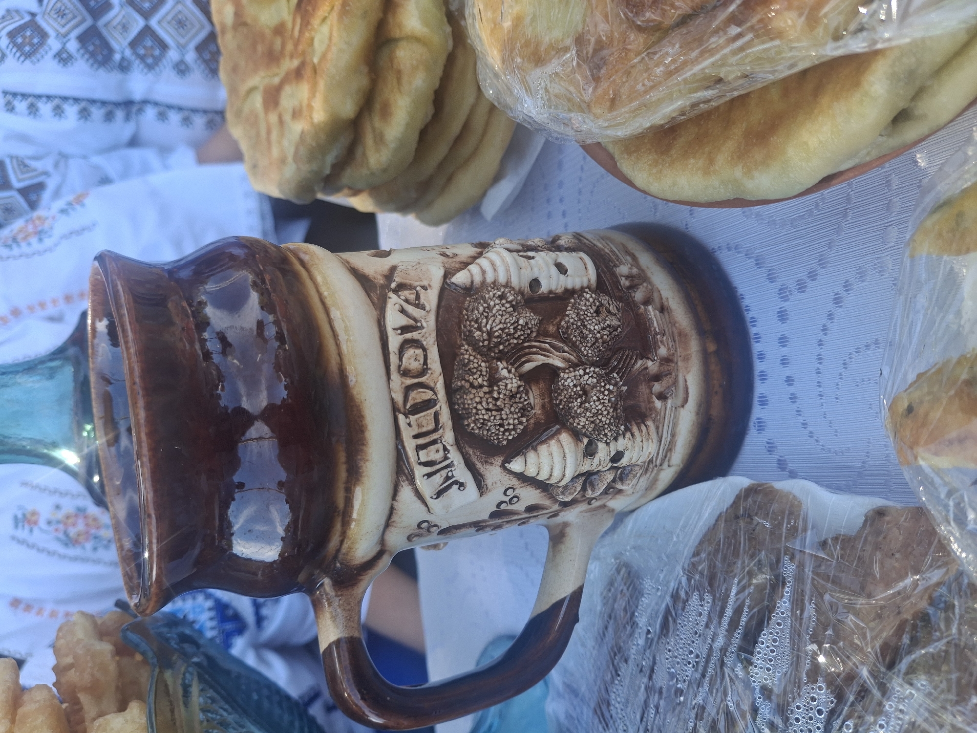 A close-up shot features a ceramic mug decorated with a relief of trees and a tower, bearing the inscription "MOLDOVA." The mug is filled with a dark liquid and sits on a white tablecloth patterned with blue and white floral designs. To the right of the mug, a stack of golden-brown, flatbread-like pastries is visible on a plate, with some pieces covered in plastic wrap. In the foreground, more pastries are wrapped in plastic, one on the left revealing a brown, dense interior, and another on the bottom right showing a lighter texture. The background is blurred, but a person wearing an embroidered white shirt is partially visible, along with a blue glass bottle. The overall impression is that of a food stall or market setting, possibly in the city of Roghi, Moldova, given the mug's inscription.