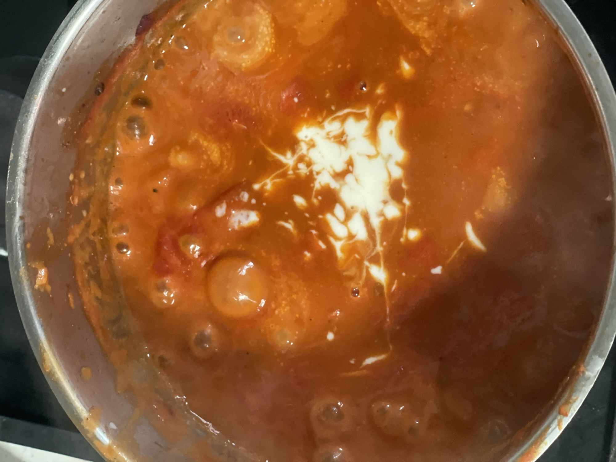 The image depicts a close-up, overhead view of a pot on a stovetop. The pot is likely made of stainless steel and contains a red, tomato-based sauce or soup. A swirl of white cream or a similar dairy product has been added to the center of the sauce, creating a marbled effect. Bubbles are visible on the surface of the sauce, indicating it is likely simmering or boiling. The scene appears to be indoors, possibly in a residential kitchen. The location in Raanana, Israel is not discernable from the image.