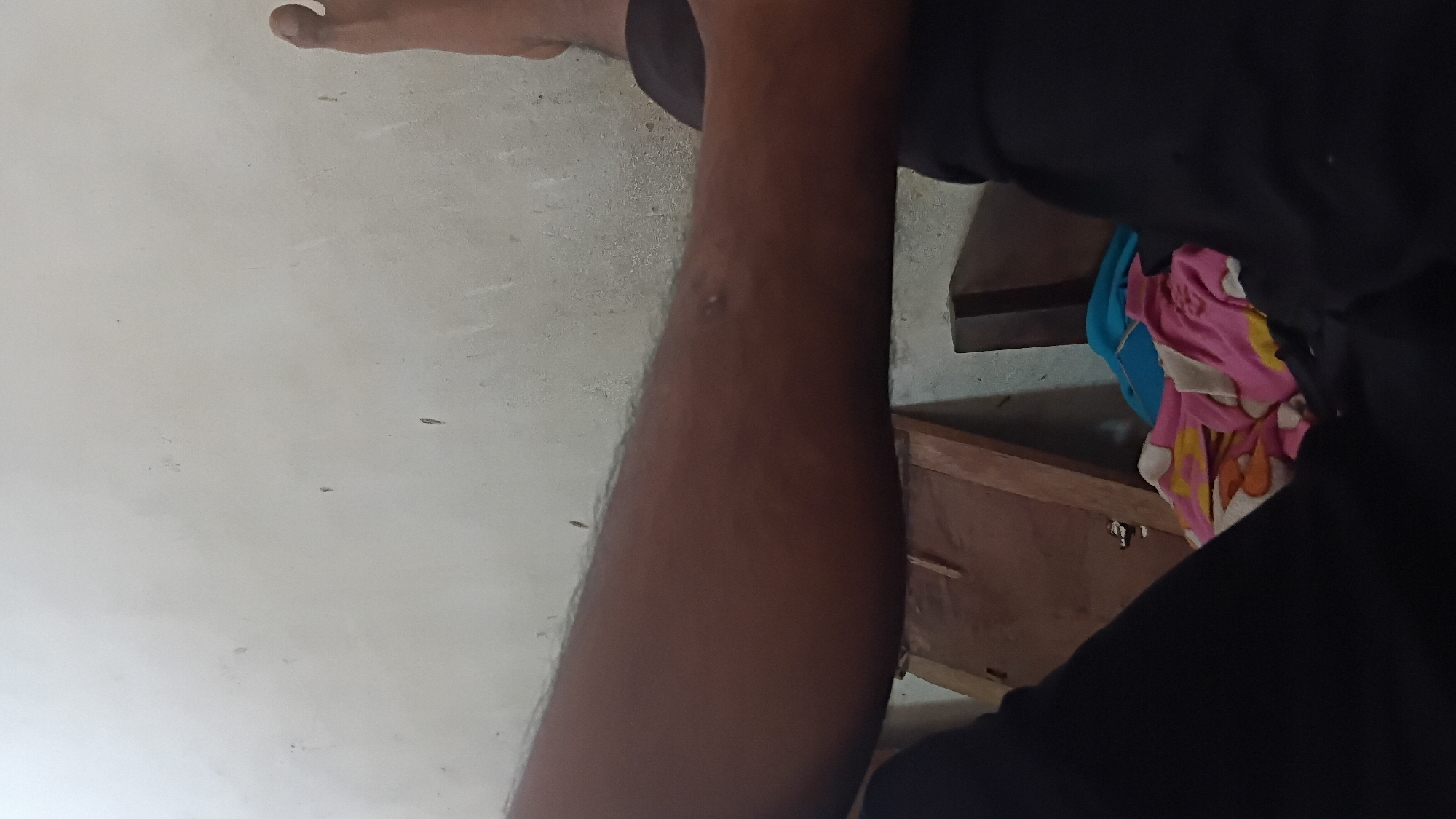 The image shows a close-up view of a person's leg and arm against a textured, off-white wall. The skin tone is dark. There are a few dark specks on the wall, consistent with dirt or imperfections. To the right of the leg, a foot is partially visible, also with dark skin. Below the arm and leg, a dark fabric, likely clothing, is visible. In the lower right portion of the image, a pile of colorful fabric, possibly blankets or clothes, is present, with patterns of pink, yellow, and white. Behind this, a wooden object is seen, possibly furniture or a wall panel, with a dark vertical element to its right. The overall impression is an indoor setting, likely a room. There is no indication of Prabumulih, Indonesia.