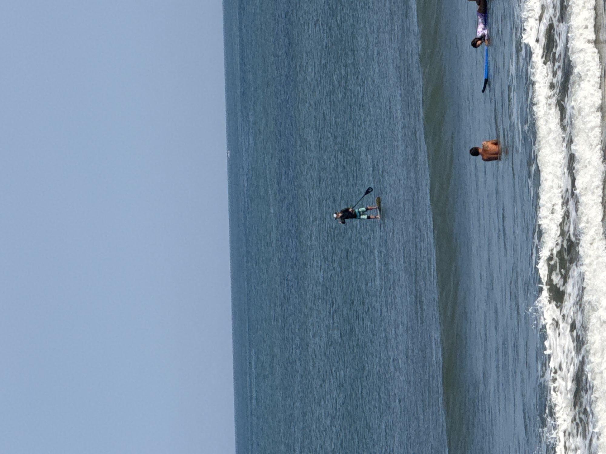 In the city of Kuta, Indonesia, an ocean scene unfolds under a clear blue sky. In the distance, a lone figure stands on a paddleboard, wielding a paddle. Closer to the shore, two individuals are visible in the water. One is a shirtless man facing away from the camera, while the other is a woman lying on a blue surfboard. White waves crash near the foreground, indicating the presence of a beach.