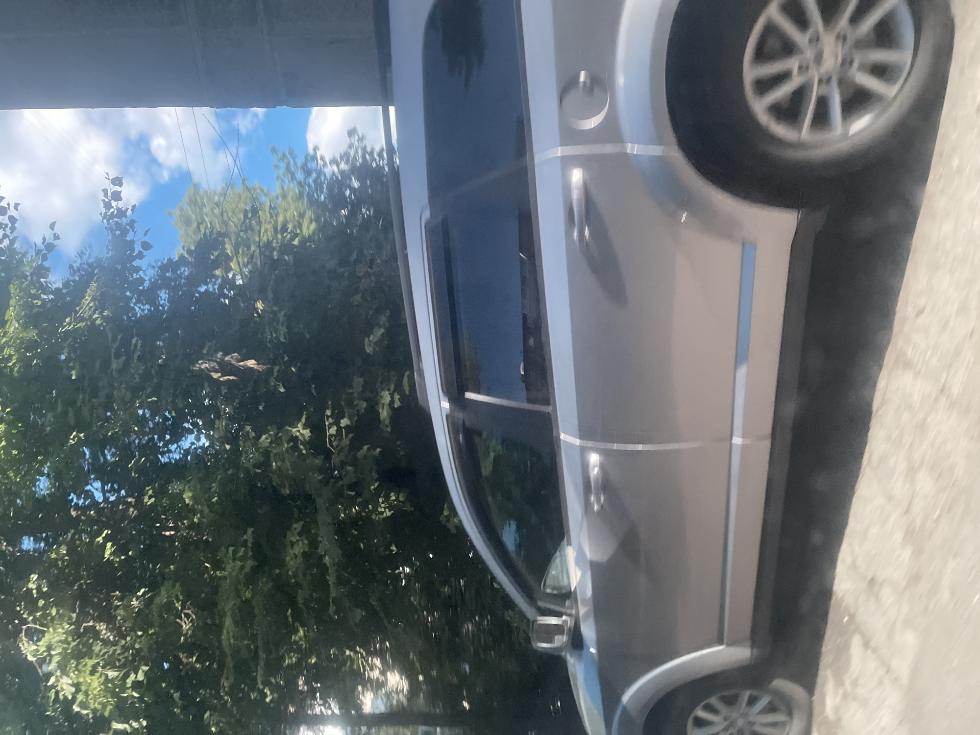 The image captures a side view of a silver SUV in Poltava, Ukraine. The vehicle appears to be in motion, as indicated by the motion blur in the foreground and on the wheels. Lush green trees with dense foliage dominate the background, interspersed with patches of blue sky and white clouds. The sky suggests it is daytime with bright sunlight, creating some glare on the car's surface and on the leaves of the trees. A portion of a grey, textured structure, possibly a building, is visible on the far right side of the frame, casting a shadow. The street surface below the car is light-colored and appears to be made of asphalt or concrete, also showing signs of motion blur. The car's windows are tinted, and a roof rack is visible. There are no people visible in the image.