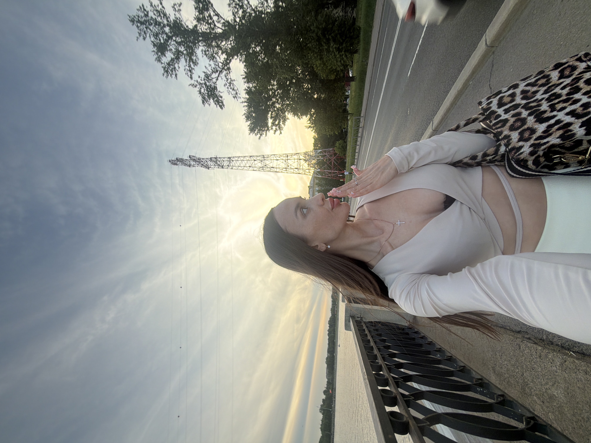 The image features a woman posing on a pedestrian walkway alongside a road and a body of water in Unknown, Russia, during what appears to be either sunrise or sunset. The woman, positioned in the foreground, has long, dark brown hair and is looking towards the upper right of the frame, her head slightly tilted back. She wears a light beige or off-white long-sleeved crop top with a deep V-neck and a cross-front design, revealing a hint of a dark undergarment and a small section of her midriff. A silver chain with a cross pendant hangs around her neck, and a small, round earring is visible in her left ear. Her left hand is raised to her mouth, with visible painted fingernails and a ring on her left ring finger, suggesting an action such as blowing a kiss or touching her lips. She carries a leopard print tote bag with black straps on her right side.
To her left, a dark, ornate metal railing separates the walkway from a calm body of water, possibly a river or canal, stretching into the distance with a tree-lined far bank. To her right, a paved road with white lane markings runs parallel to the walkway. A blurred, light-colored vehicle is in motion on the road, moving from right to left. Beyond the road, a grassy area features several mature trees, and a tall, prominent electricity transmission tower (pylon) with a lattice structure rises against the sky. The sky itself displays a gradient of soft blue at the top transitioning to warm yellow and orange hues near the horizon, indicative of golden hour light. Wispy, streaky clouds are visible, and several power lines with circular markers traverse the upper portion of the sky.