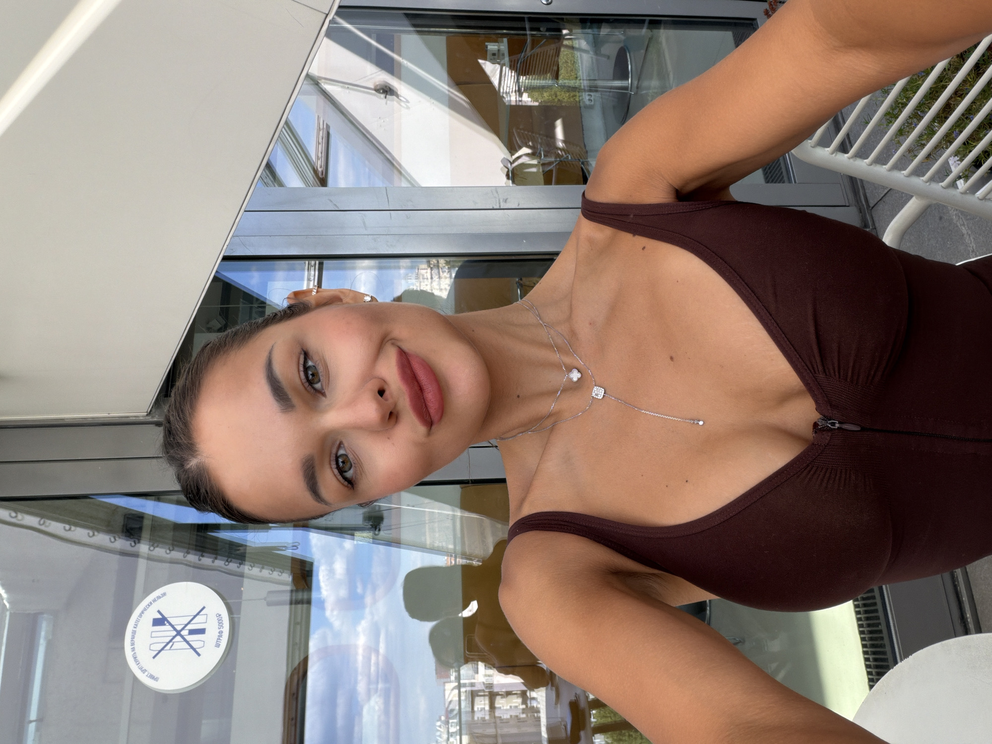 A young woman is captured in a headshot selfie, looking directly at the camera with a subtle smile. The image is oriented sideways. She has dark hair pulled back, strong brows, and is wearing visible makeup including eyeliner and a nude-pink lipstick. She is wearing a dark brown, possibly ribbed, sleeveless top or dress with thin straps and a zipper visible on the chest. A delicate silver necklace with two small pendants, one appearing as a four-leaf clover and the other a square with a gem, extends down her décolletage.
The background consists of large glass windows or doors, through which reflections of an urban environment are visible. These reflections show parts of a building's structure, clear blue skies with some clouds, and what appears to be a white slatted chair. The bright natural light suggests it is daytime.
A white circular sticker is affixed to the glass behind her. The text on the sticker, written in Cyrillic, reads "НАВЕРХЕ КАТЕГОРИЧЕСКИ НЕЛЬЗЯ" (Absolutely forbidden upstairs), below which is an icon of crossed-out items (possibly glasses and a bottle, indicating no food or drinks). To the right, "ШТРАФ 5000Р" (Fine 5000R) is visible, strongly indicating a location in Russia, consistent with the given context of Moscow.