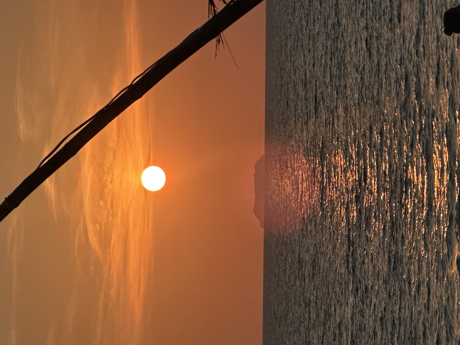 This image depicts a sunset over the sea, located in Phra Ae, Thailand. The sky is predominantly orange-red with subtle, elongated cloud formations. A bright, circular white sun disc is positioned in the upper-left quadrant, casting a prominent vertical reflection path across the dark, wavy ocean surface below. A low, dark, hazy island silhouette rests on the horizon, slightly right of the frame's horizontal center. In the foreground, a thick, dark object, consistent with a diagonal pole or tree trunk, extends from the upper-right corner downwards and towards the left. Fine, dark linear elements are visible along its surface, and dark, spiky fronds are partially visible at the bottom-right of the frame, emanating from or near the diagonal object. No human subjects or discernible actions are present.