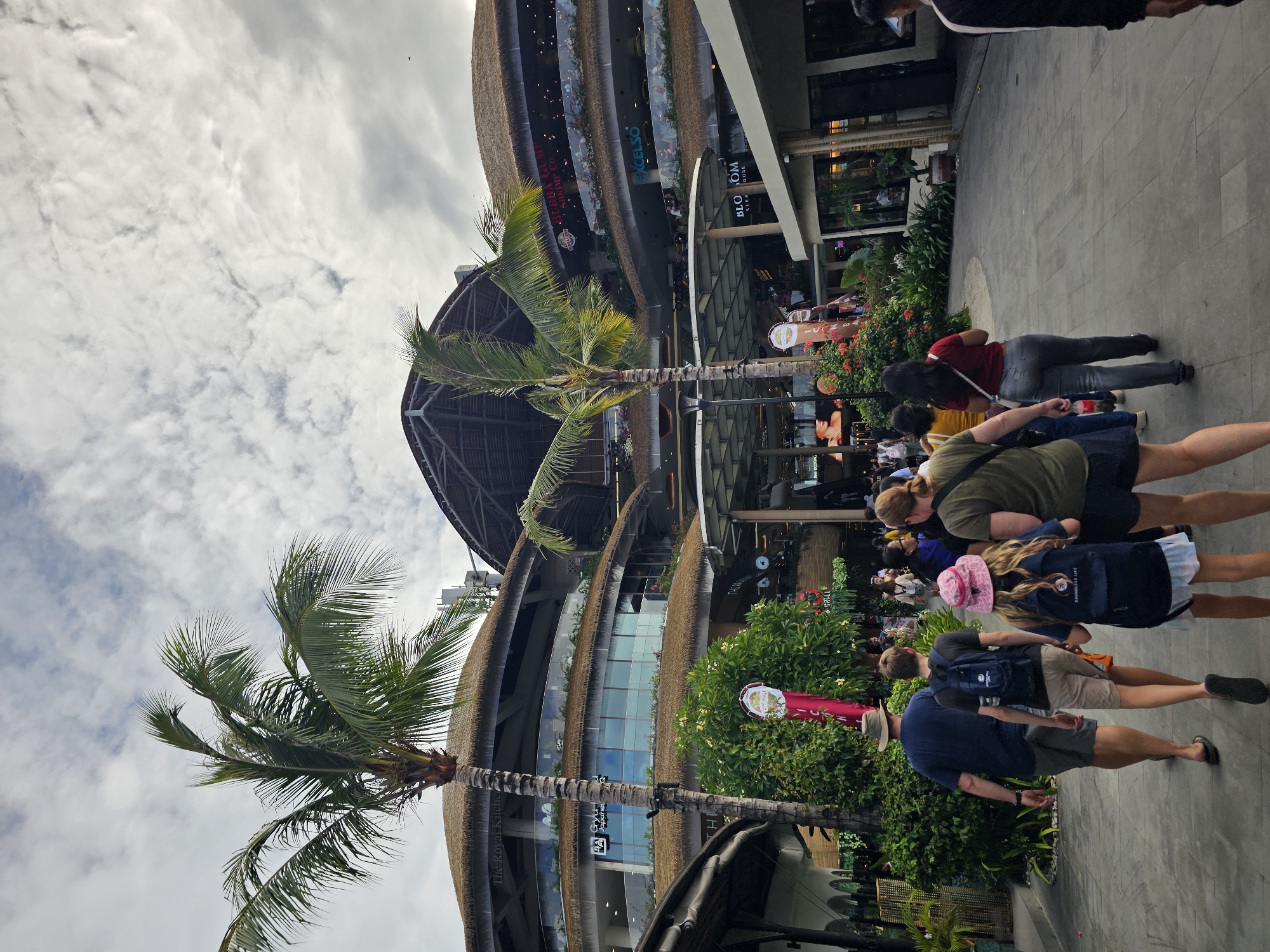 In Denpasar, Indonesia, a group of people are walking in a modern outdoor shopping mall. The mall features multi-level architecture with thatched roofs and glass railings. Palm trees are interspersed throughout the complex, adding to the tropical ambiance. The sky is cloudy. In the foreground, a family walks away from the camera. The father, wearing a blue t-shirt and hat, walks on the left. Next to him, a young boy in a black t-shirt and backpack follows. On the right, a young girl with a pink hat and backpack holds hands with a woman in a green shirt. Other shoppers can be seen in the background, some walking and some browsing the storefronts. Store names like "The Royal Kitchen," "Gyuhan Japanese," "Bubba Gump Shrimp Co.," "Excelso," and "Blossom" are visible on the buildings, indicating a diverse range of dining and retail options. The ground is paved with grey tiles, and lush green plants line the pathways.