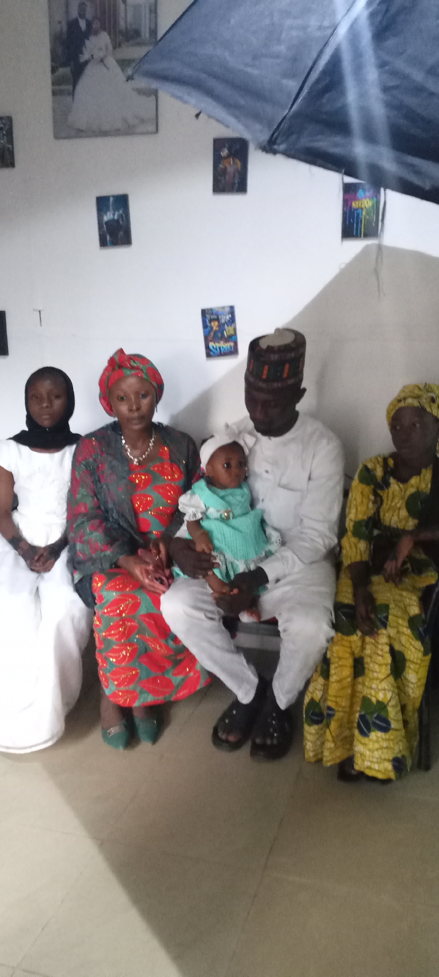 The image depicts a group of five people indoors. From left to right, there is a young woman wearing a white dress and black headscarf, a woman in a red and green patterned dress and matching headscarf with a grey shawl, a man in a white outfit and patterned cap holding a baby in a light blue dress, and a woman in a yellow patterned dress and headscarf. The group is seated, posing for the photograph.
The background features a white wall decorated with various pictures, including a wedding photograph. The floor is a light, neutral color. A large, dark object, possibly part of a photography setup, is visible in the upper right corner.