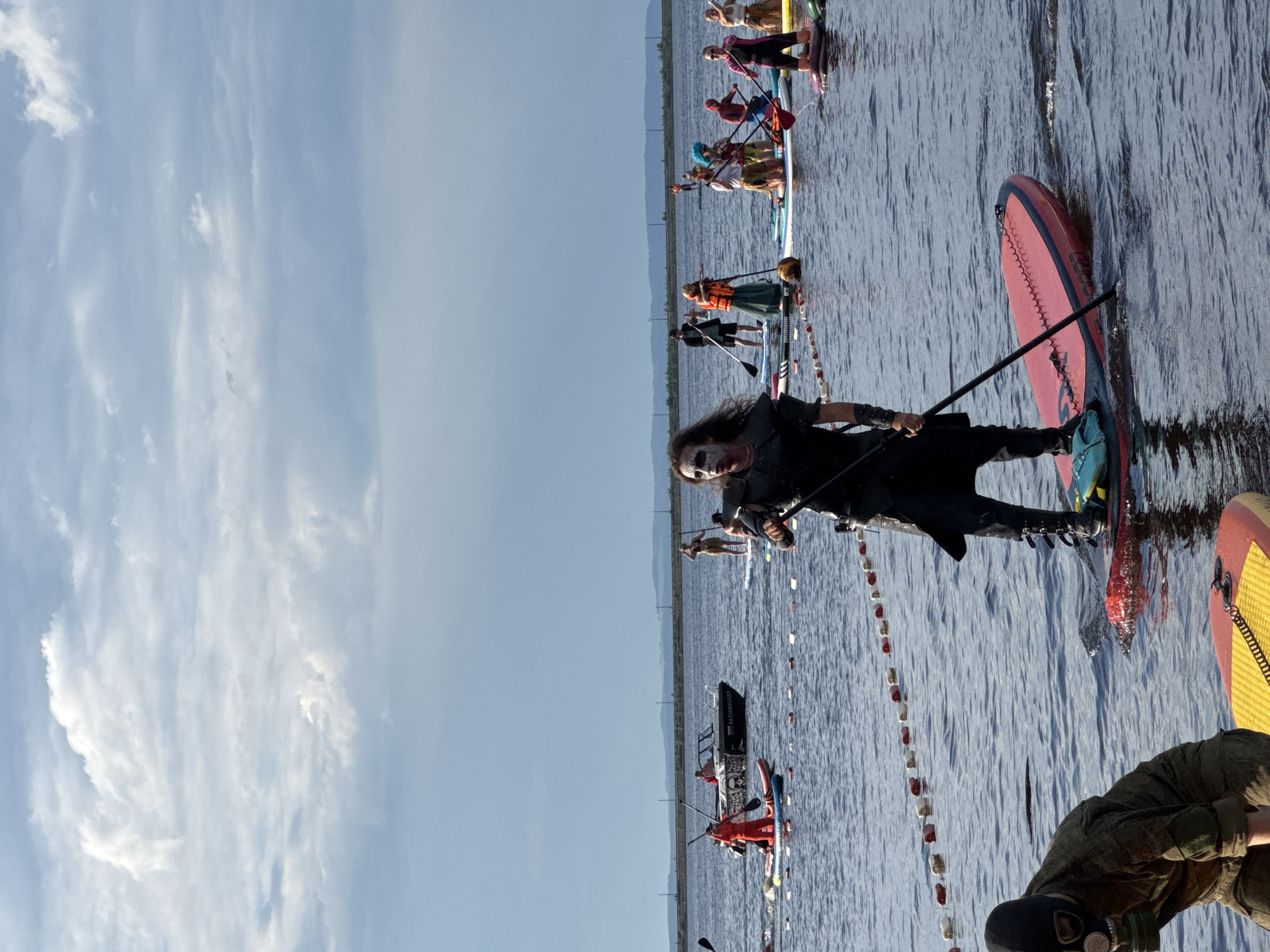 In Chaikovsky, Russia, on the Kama River, a group of individuals participates in a stand-up paddleboarding event. The main subject, in the foreground and facing the camera, is dressed in a dark, elaborate costume with white face paint and dark eye makeup, holding a paddle. Behind them, several other participants are visible on paddleboards, some in costumes ranging from a gas mask and military-style attire to a Spider-Man suit and other colorful outfits. A small motorboat is present in the middle ground, and a marker line with buoys stretches across the water. The environment is a wide, calm body of water under a cloudy sky, with a distant shoreline featuring trees and buildings. The event appears to be a recreational or themed gathering taking place on a sunny day with overcast clouds.