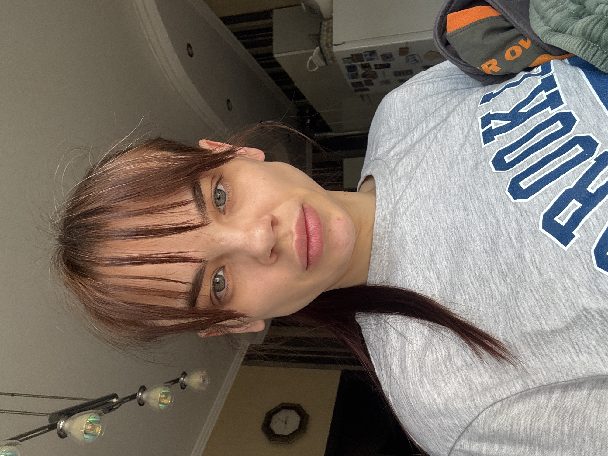A woman with auburn hair and bangs, wearing a gray t-shirt with blue lettering, possibly "BROOKLYN," is shown indoors. Background details include a ceiling light fixture, a clock on the wall, and glimpses of kitchen appliances. The location in Rostov-on-Don, Russia, is not discernible from the image.