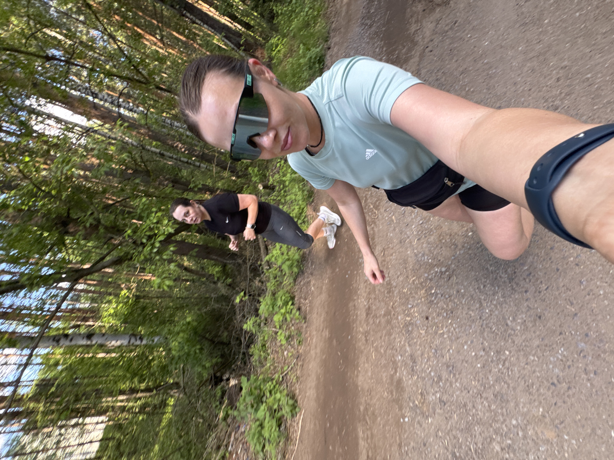 The image shows two women running on a dirt path surrounded by a forest. The woman closer to the camera is wearing sunglasses, a light blue t-shirt with an Adidas logo, black shorts, and a fitness tracker on her wrist. The other woman is further down the path wearing a black t-shirt and gray running tights, and white sneakers. The path is surrounded by green foliage and trees, with dappled sunlight filtering through the canopy. It is understood that the location is the city of Chaikovsky, Russia.